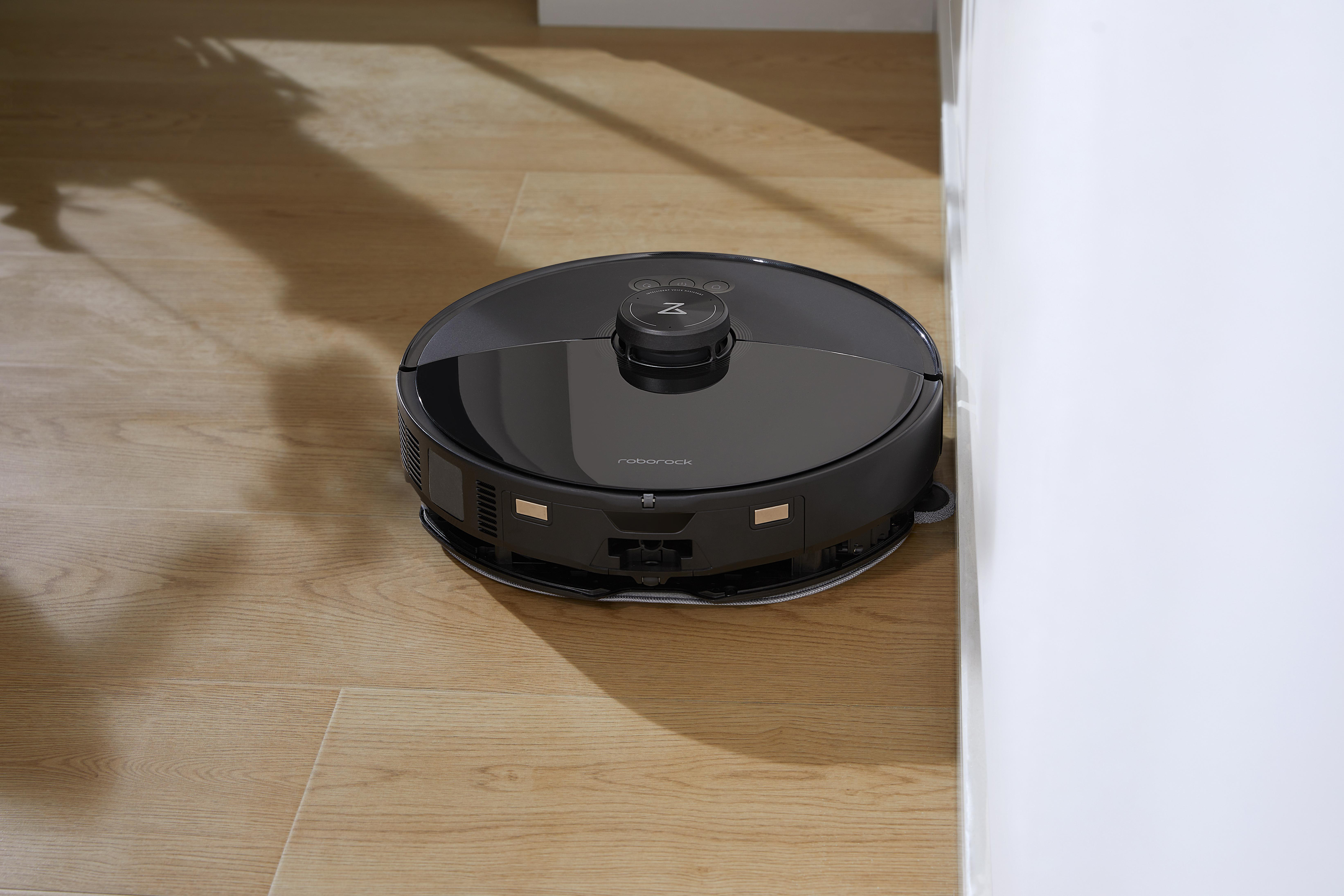 Ein schwarzer Roboter-Staubsauger steht auf einem Holzboden neben einer weißen Wand, eine Ecke ist sichtbar.