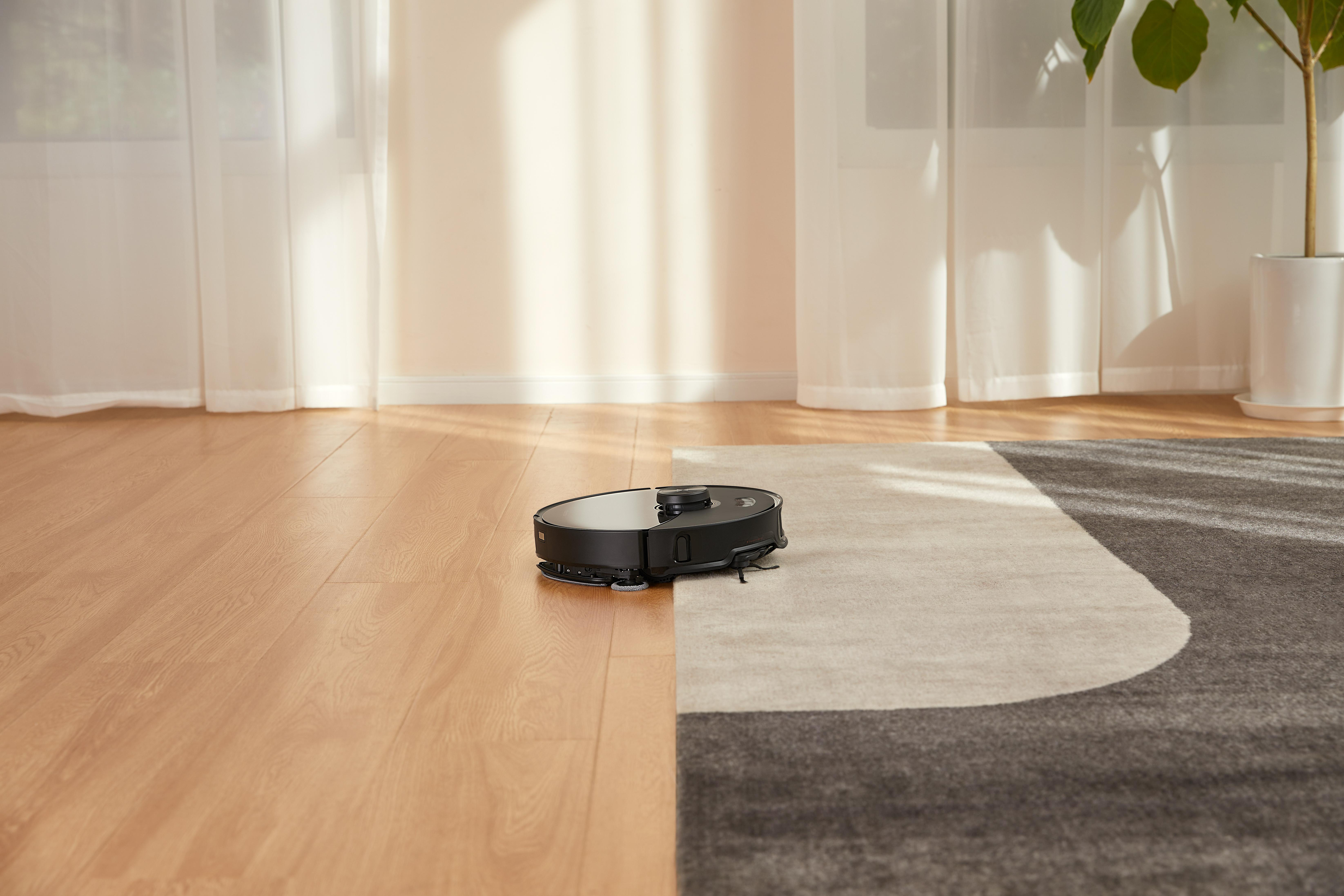 Ein schwarzer Roboter-Staubsauger reinigt einen Raum mit Holzboden und Teppich.
