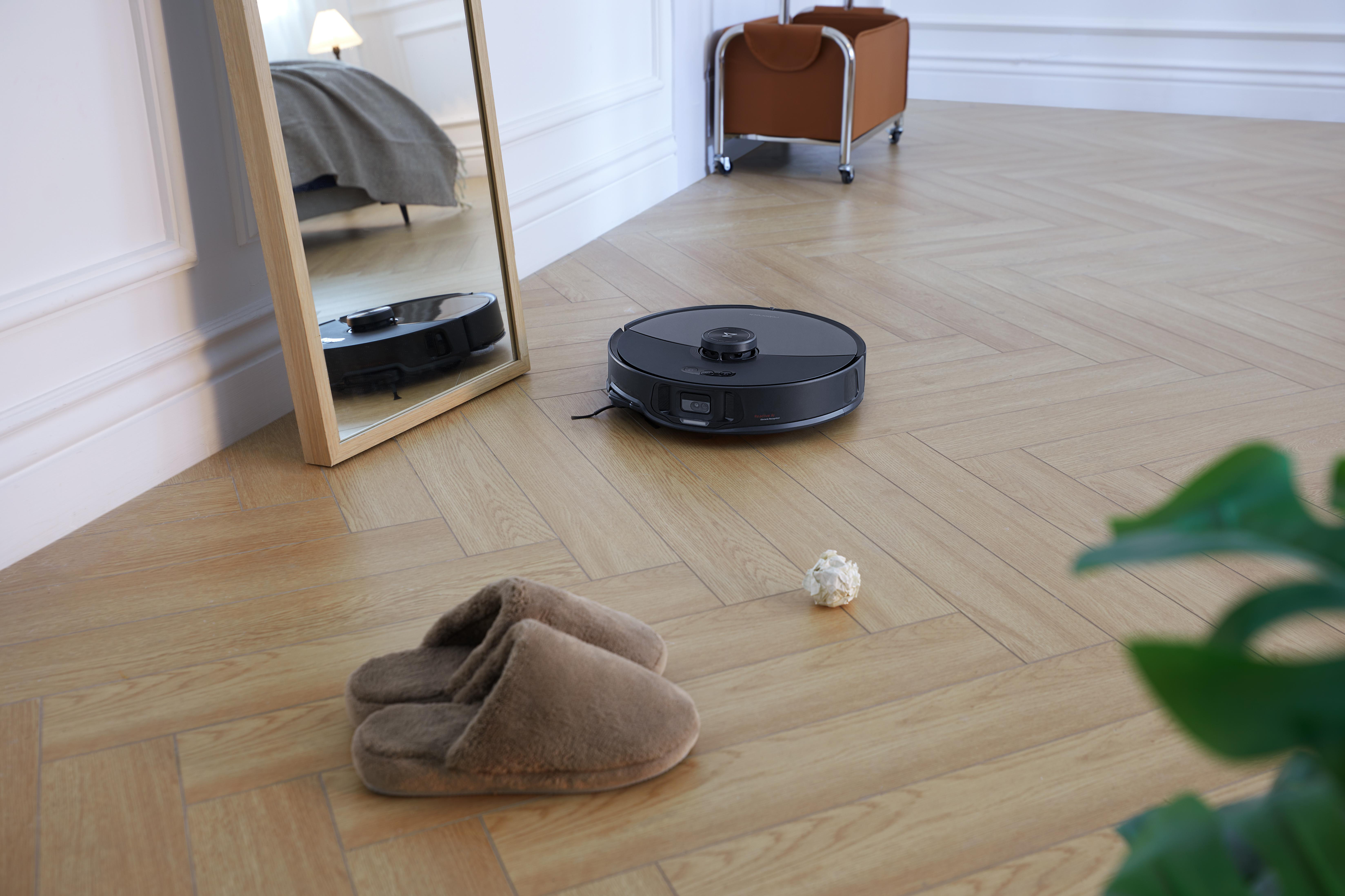 Ein Roboter-Staubsauger auf Holzboden neben Hausschuhen und einem Spiegel. Der Spiegel spiegelt den Staubsauger.
