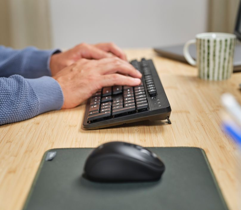 Manos tecleando en un teclado negro, con un ratón y una taza.