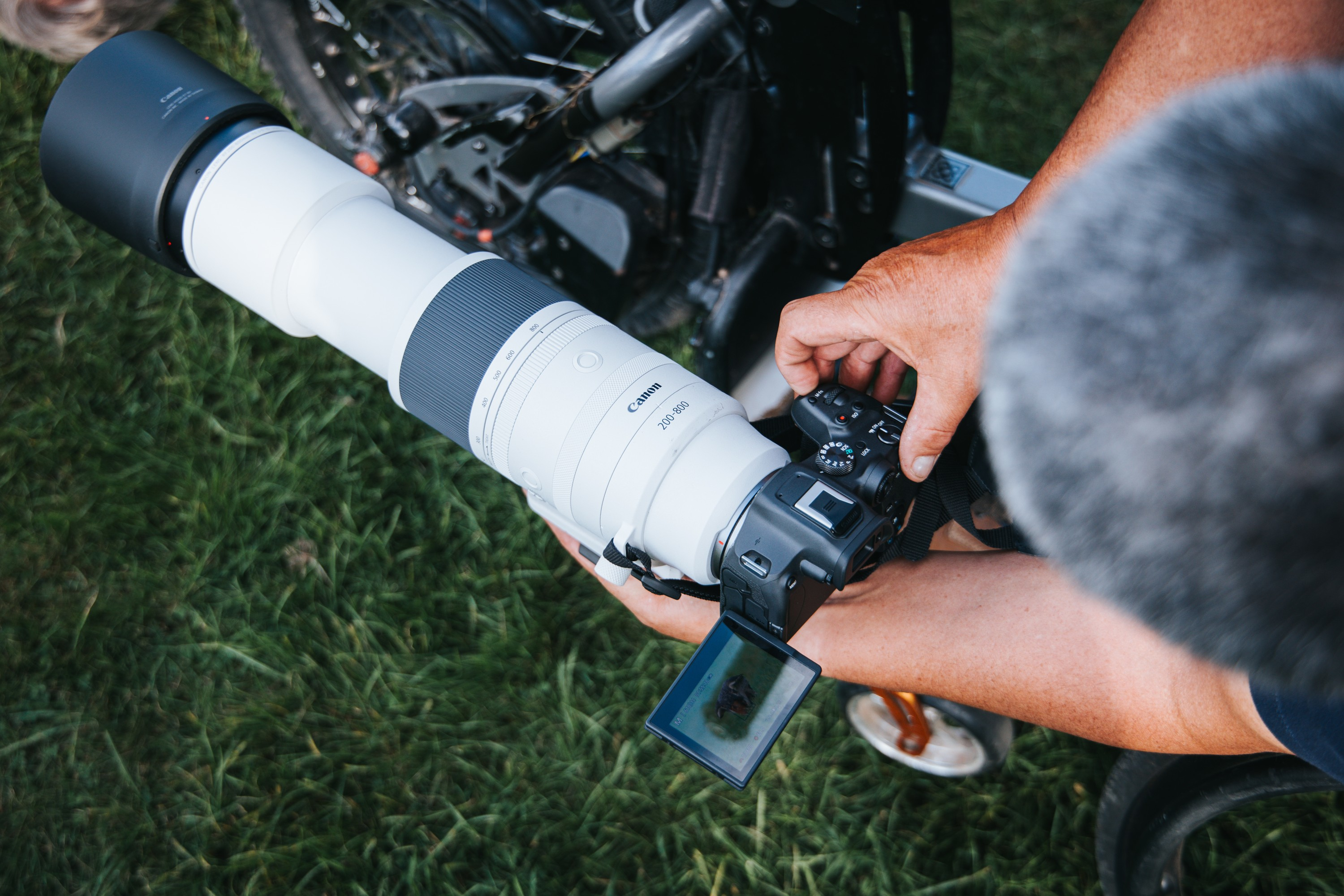Person hält eine Kamera mit einem großen weißen Objektiv. Der Hintergrund ist grünes Gras und ein Fahrrad.