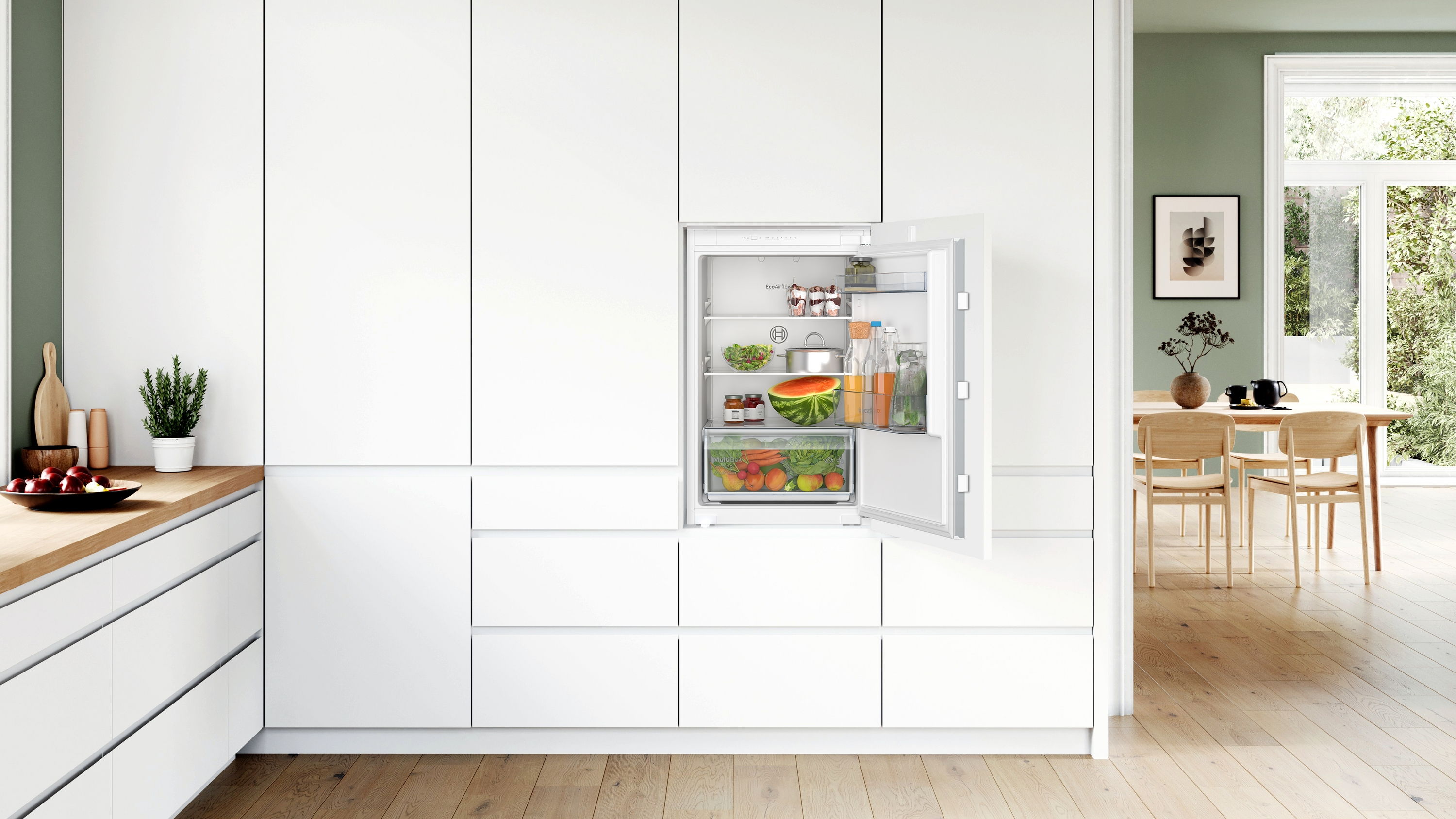 Eine Küche mit geöffnetem Kühlschrank, der Lebensmittel zeigt. Weiße Schränke und eine Holz-Arbeitsplatte sind sichtbar.