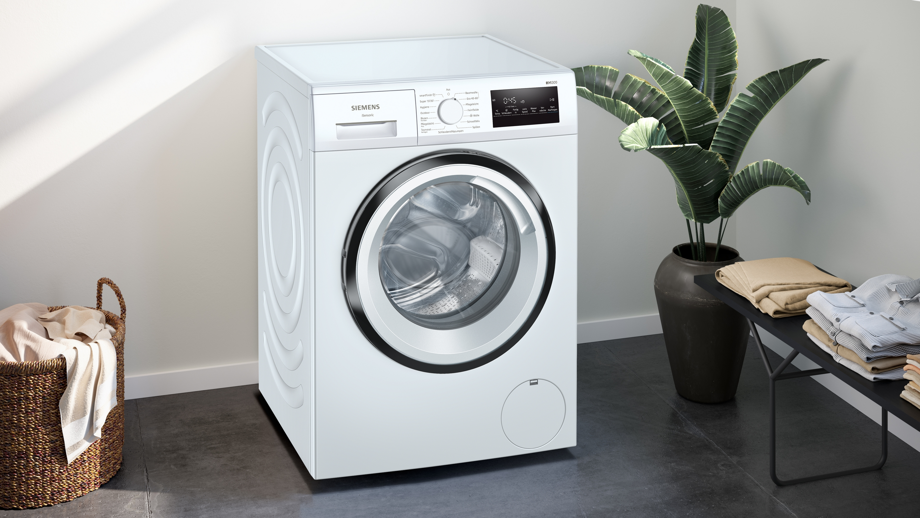 Weiße Waschmaschine mit Bedienfeld und Wäschekorb. Pflanzen im Hintergrund.