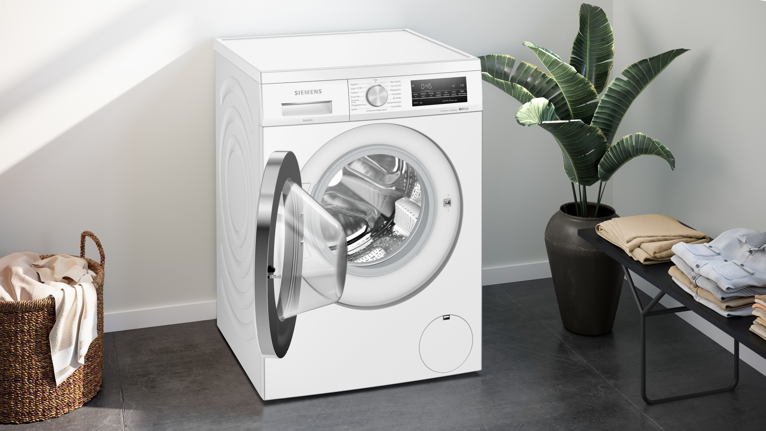 Weiße Waschmaschine mit offener Tür, neben Pflanze und Wäschekorb.