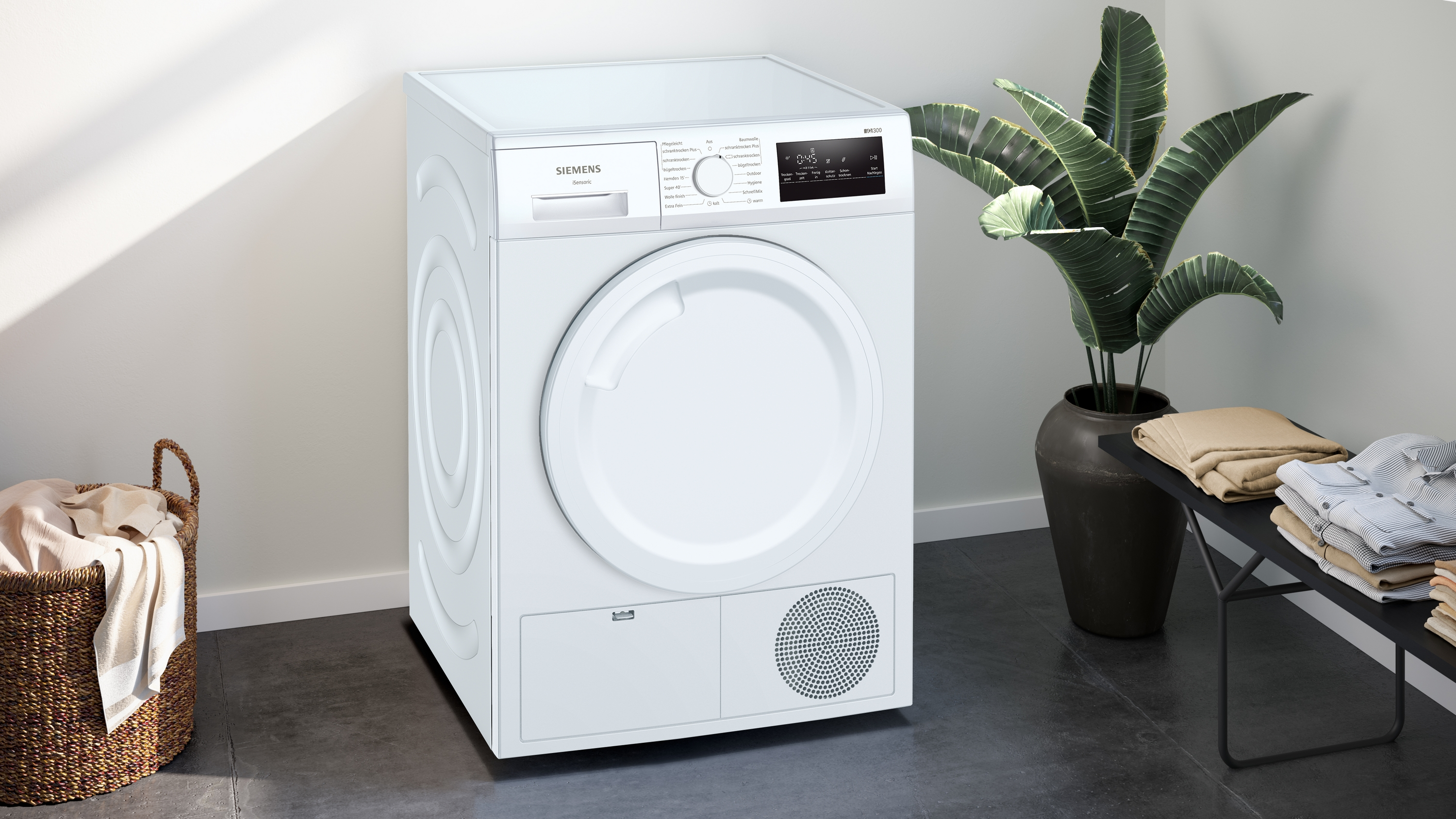 Weißer Trockner in einem Raum. Wäschekorb und eine Pflanze daneben. Sauberes Design.