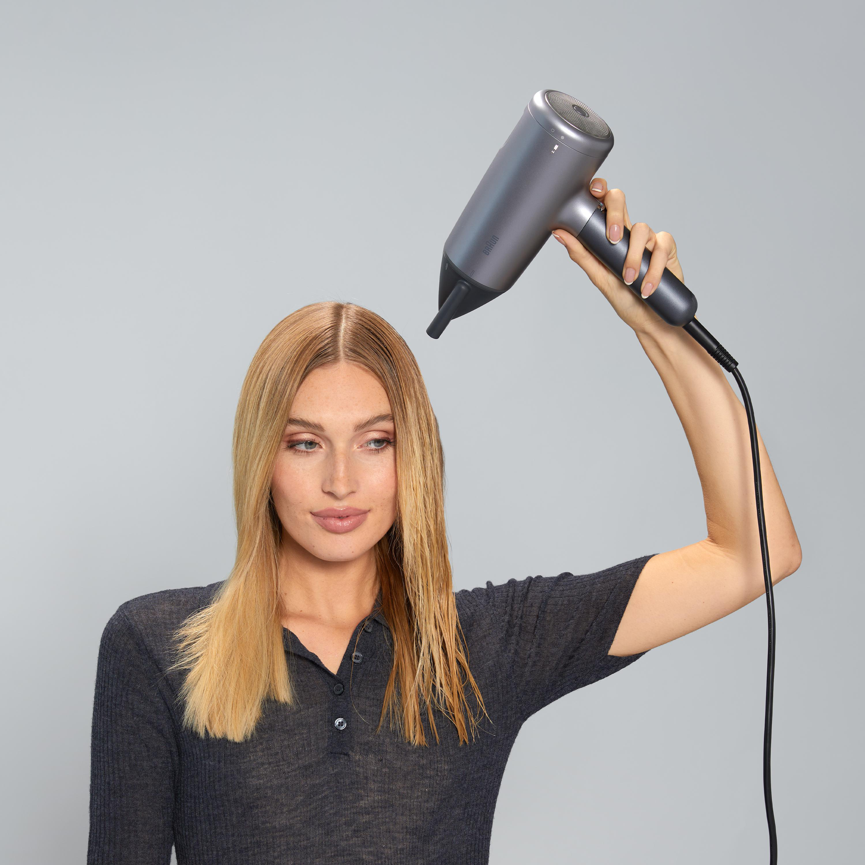 Frau benutzt grauen Haartrockner. Blondes Haar ist herunterhängend, der Trockner ist darauf gerichtet.