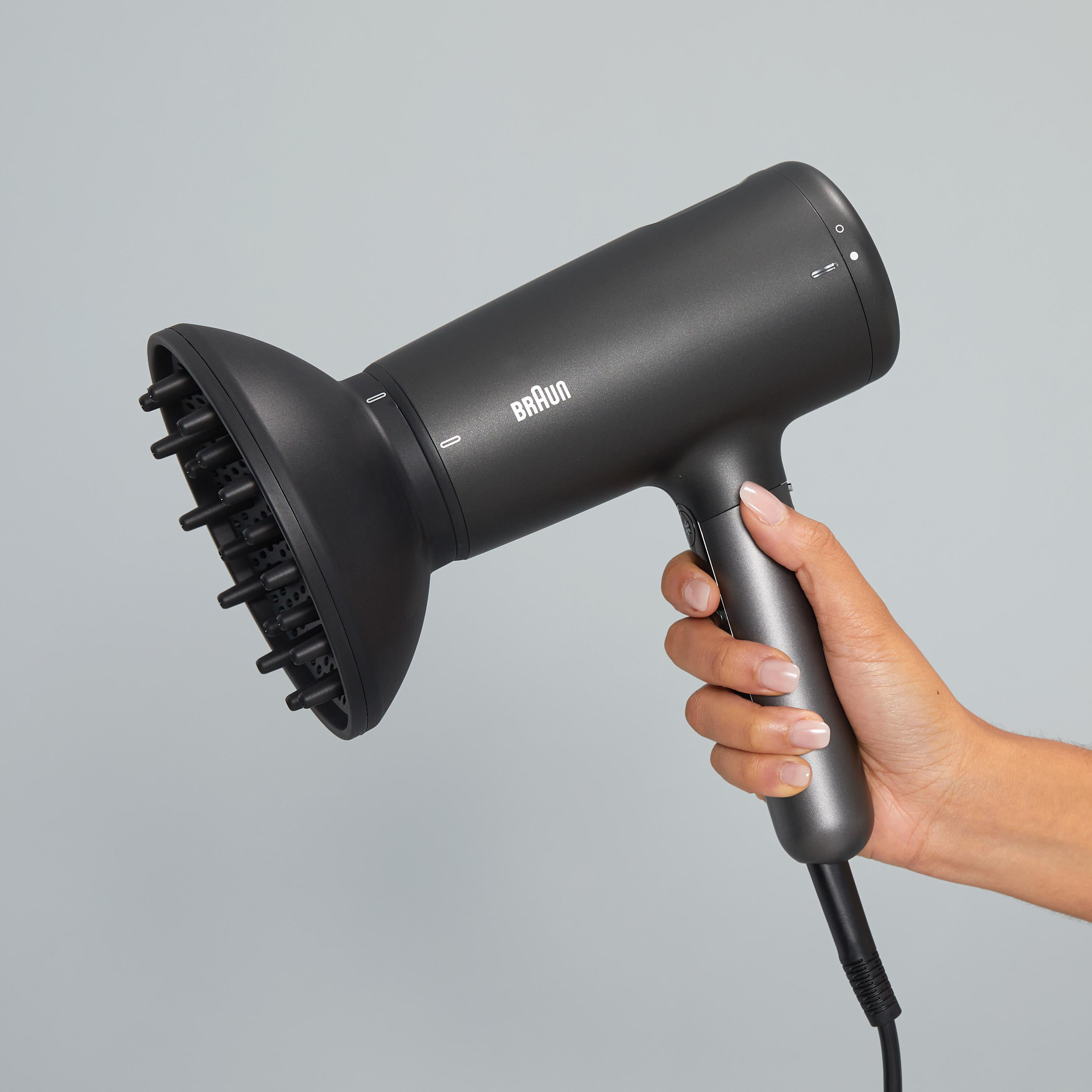 Ein schwarzer Braun-Haartrockner wird von einer Hand vor grauem Hintergrund gehalten.