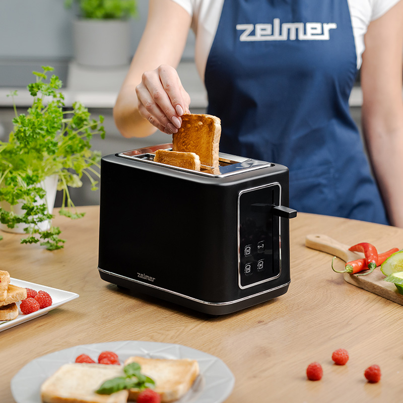 Osoba wkładająca tosty do czarnego tostera. Widoczne inne produkty spożywcze.