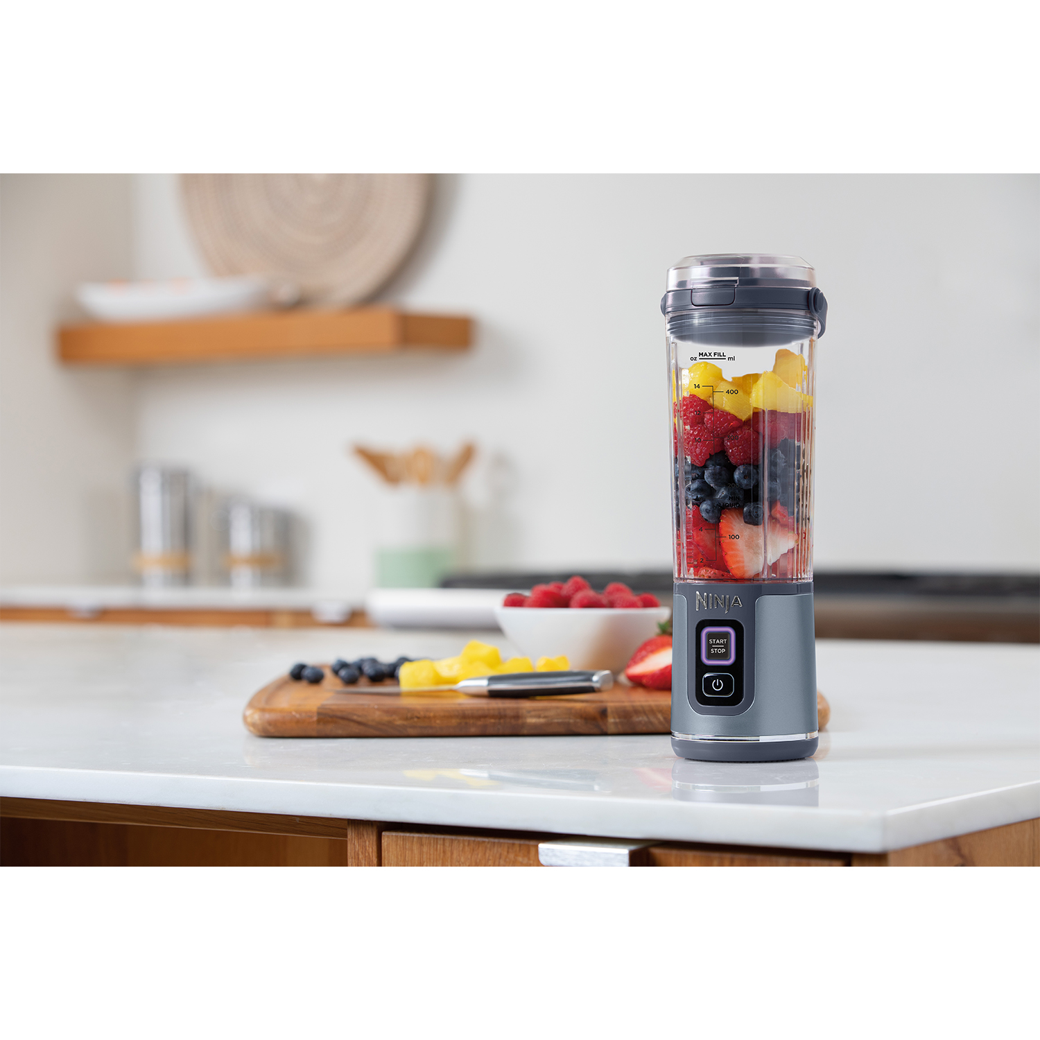 Frullatore grigio con contenitore pieno di frutta. Su un piano di lavoro bianco con tagliere.