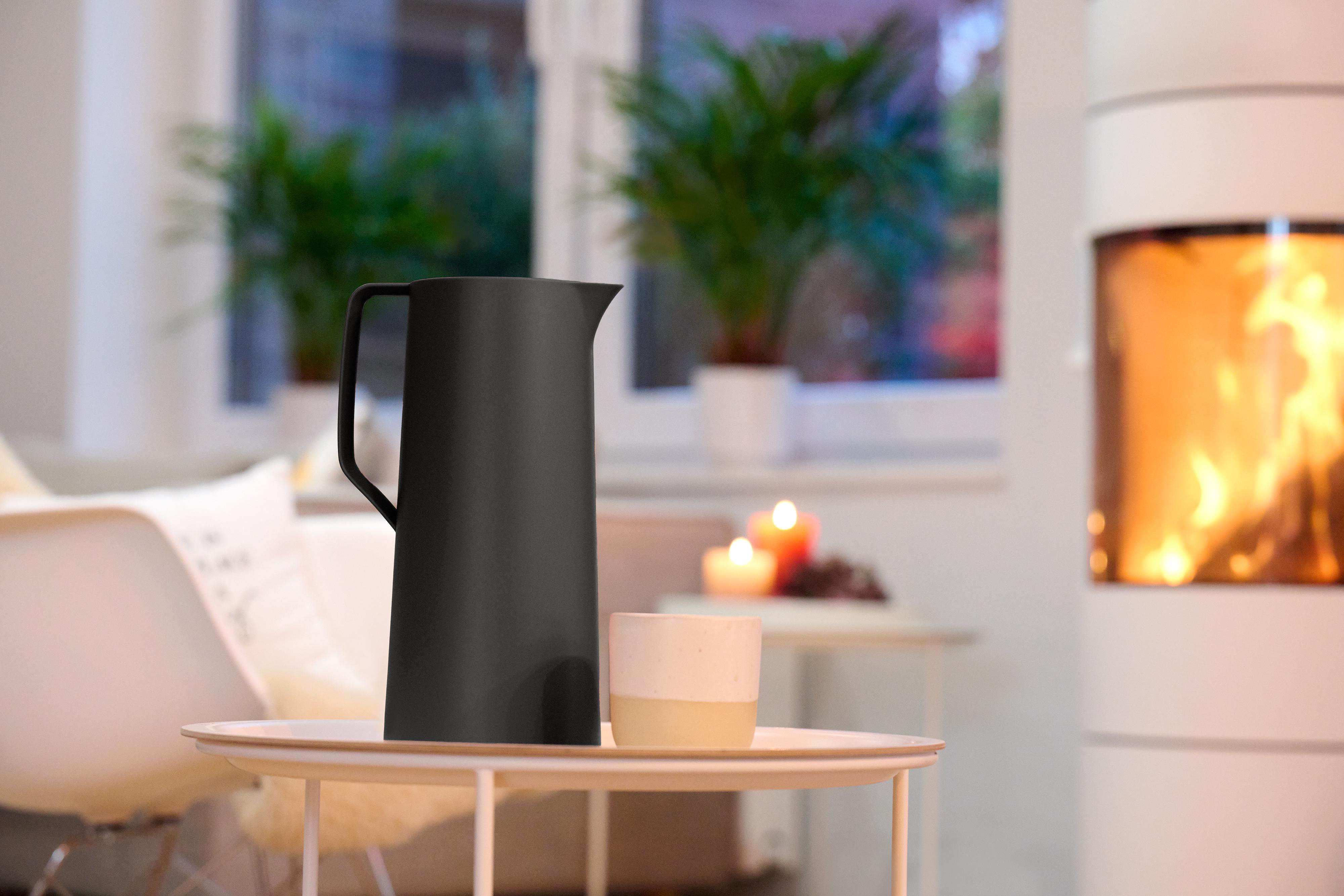 Schwarzer Krug und beigefarbene Tasse auf einem Tisch. Kamin im Hintergrund, mit Pflanzen und Kerzen.
