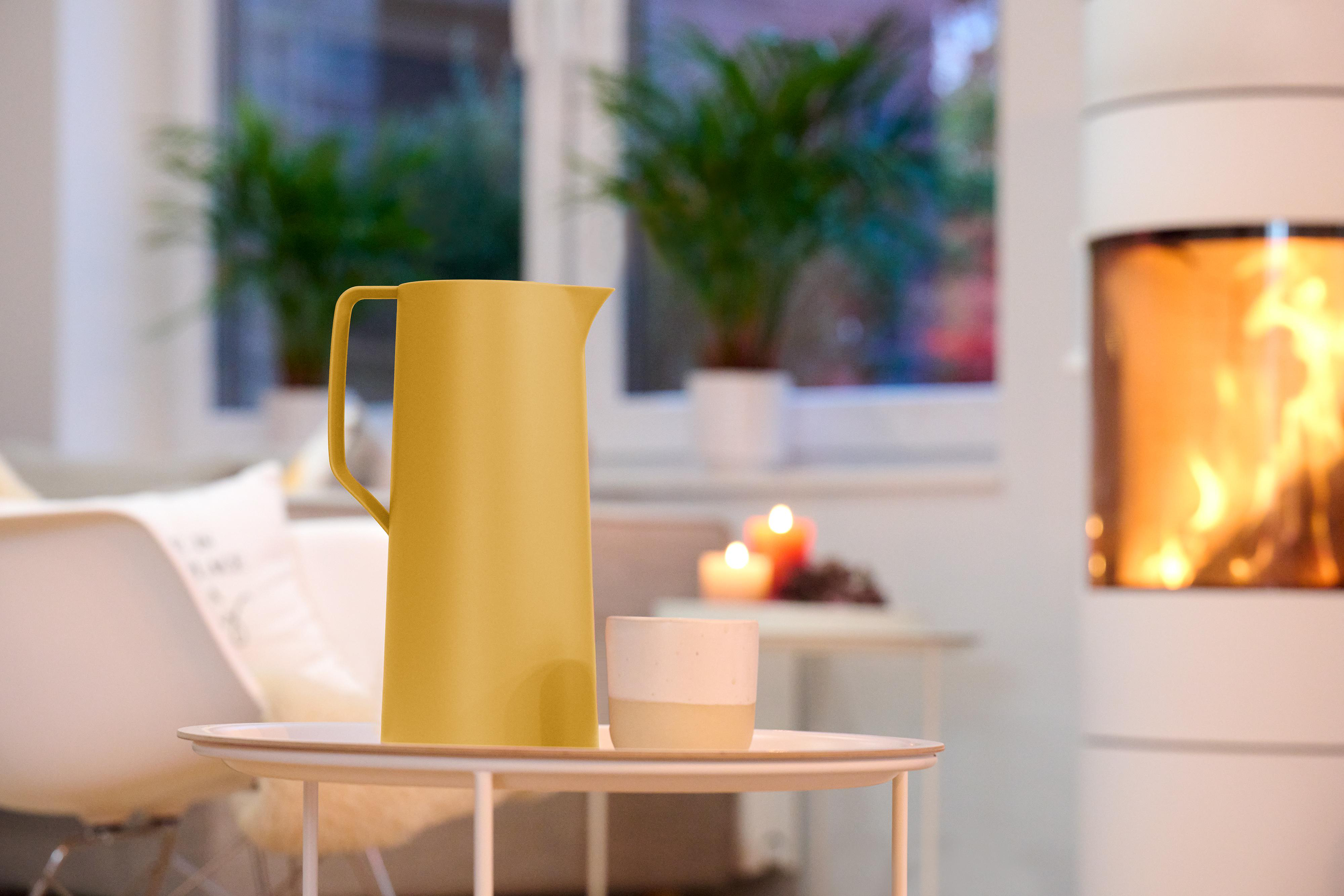Ein gelber Krug und eine weiße Tasse auf einem Tisch. Ein Kamin und Pflanzen im Hintergrund.