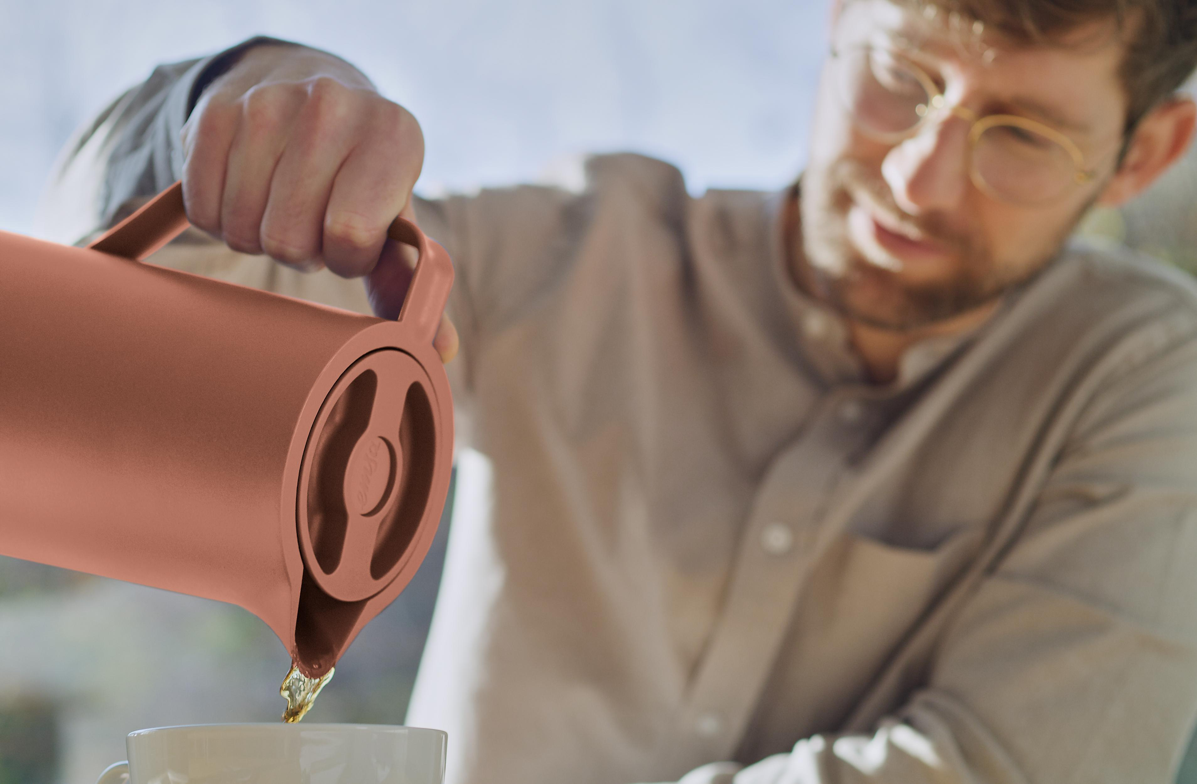Eine Person gießt Flüssigkeit aus einer kupferfarbenen Thermoskanne in eine weiße Tasse. Unscharfer Hintergrund.