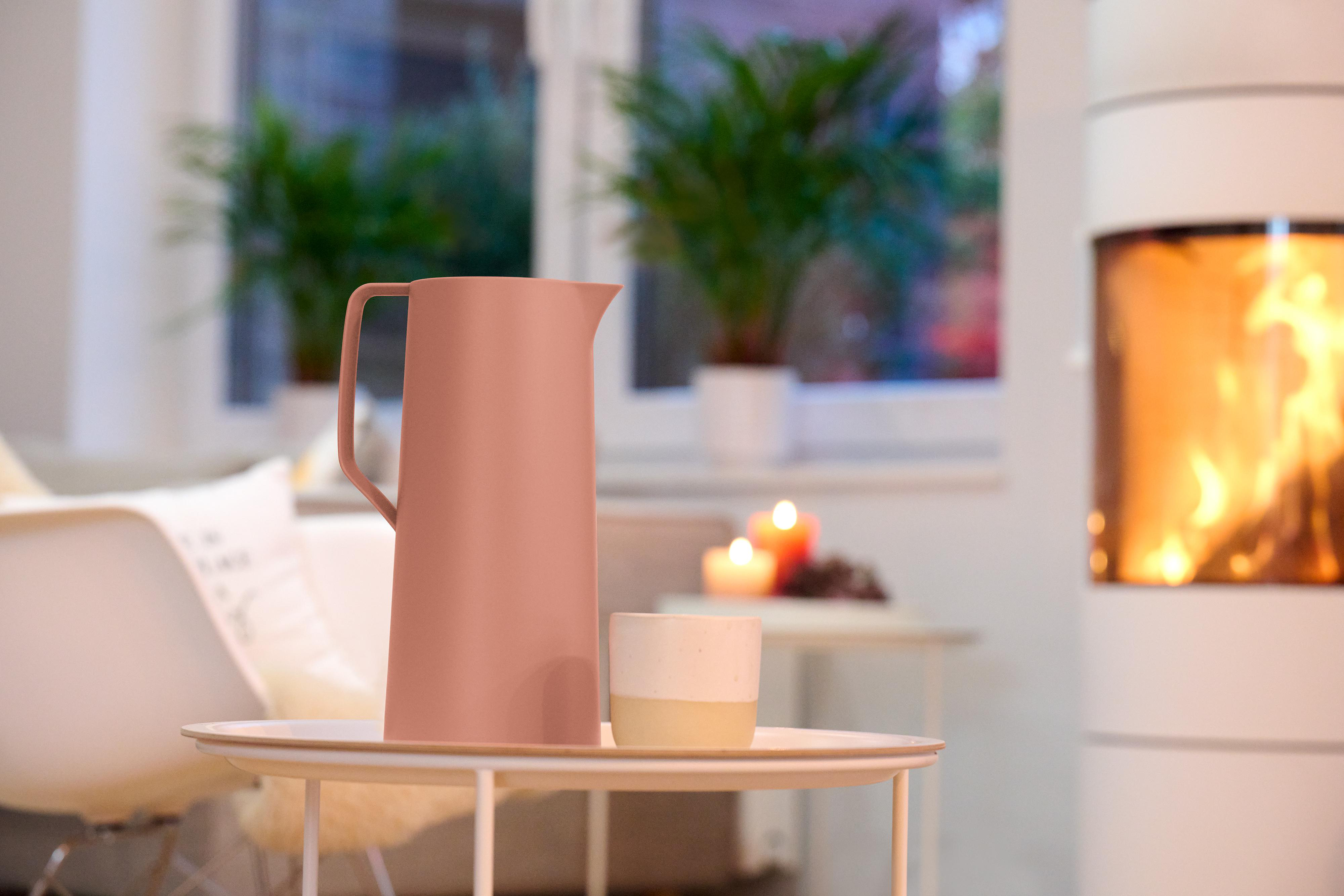 Rosa Wasserkrug und weiße Tasse auf einem Tisch. Pflanzen und Kamin im Hintergrund.