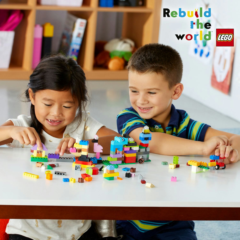 Zwei Kinder bauen mit bunten Lego-Steinen auf einem weißen Tisch, das Lego-Logo ist zu sehen.