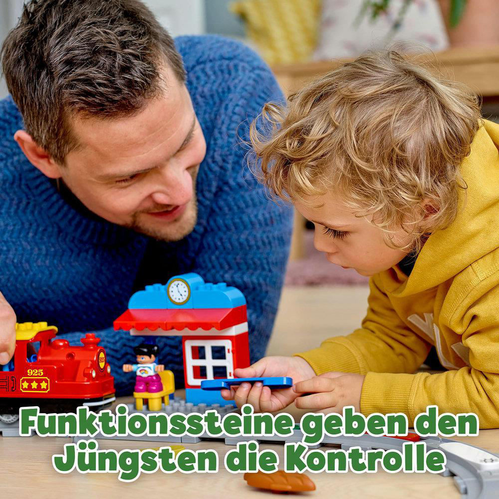 Ein Vater und sein Sohn spielen mit LEGO Duplo-Steinen, mit einem roten Zug und einem Gebäude mit Uhr.