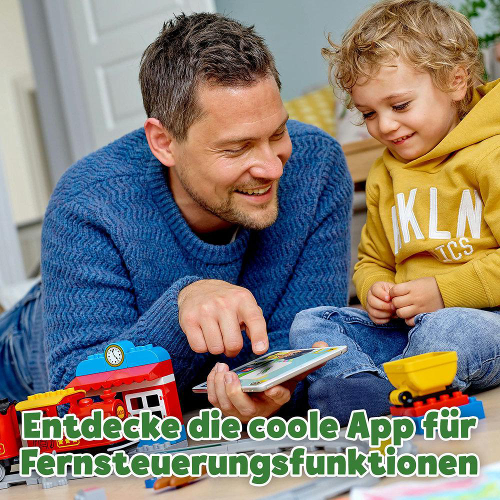 Ein Vater und Sohn spielen mit einem Tablet und einem Spielzeugeisenbahnset auf dem Boden, lächelnd.
