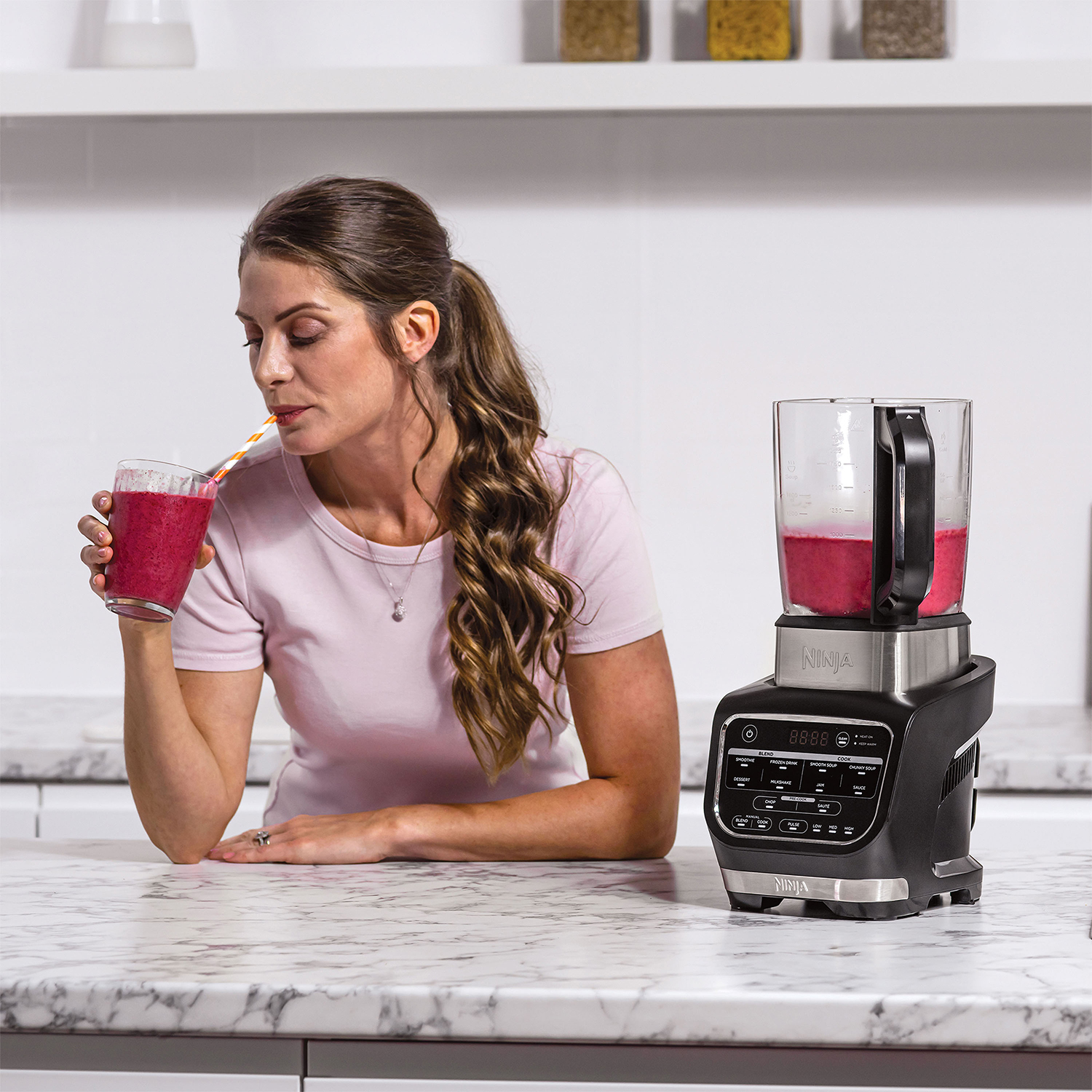 Una donna beve da un bicchiere con una cannuccia, accanto a un frullatore nero su un piano di lavoro bianco.