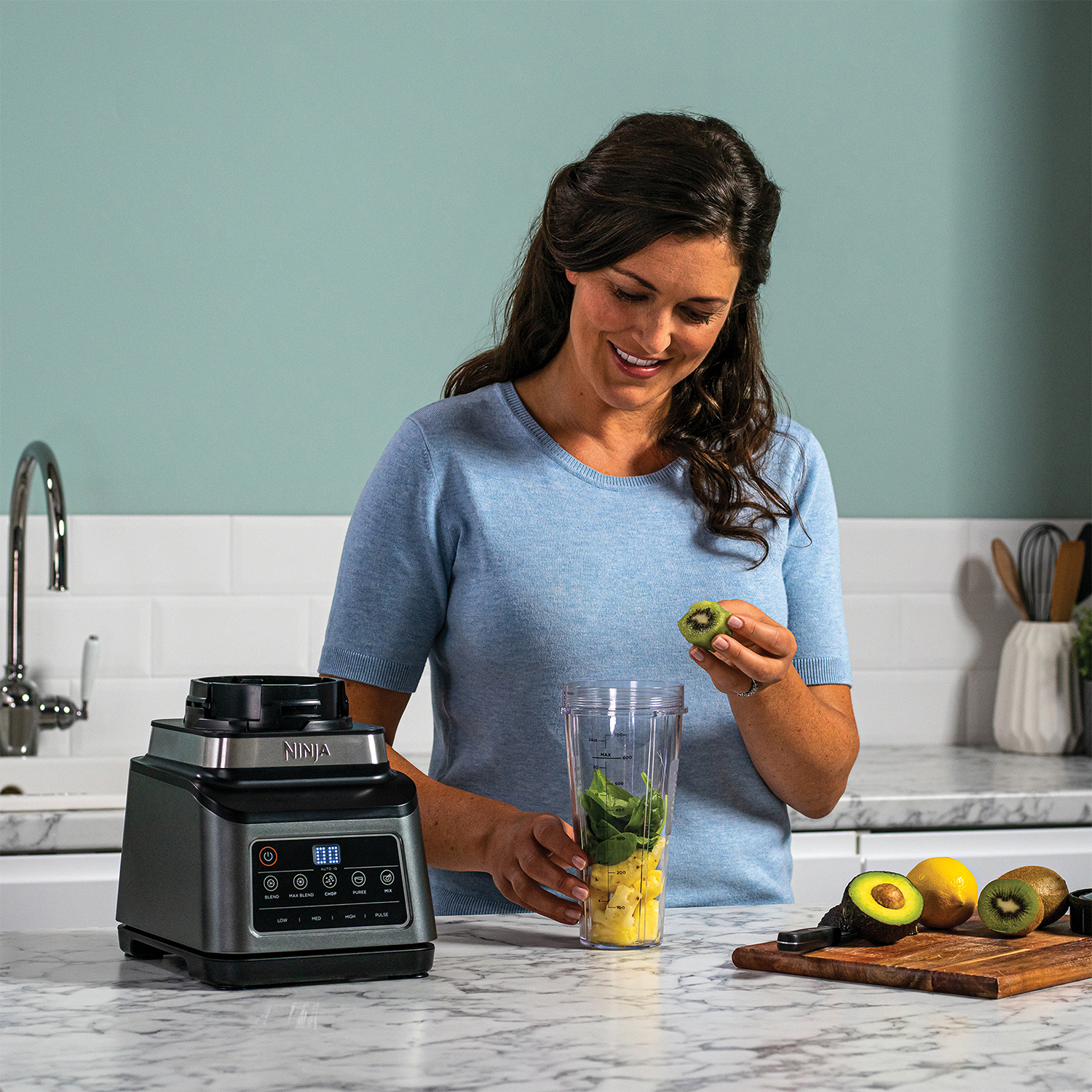 Donna che prepara un frullato. Tiene un kiwi e prepara gli ingredienti in un frullatore.