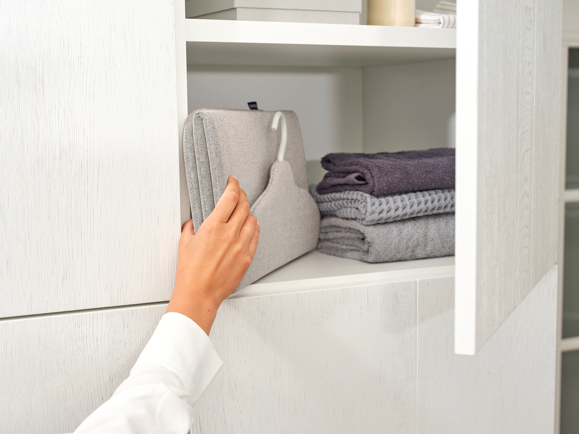 Eine Hand greift in einen weißen Schrank, um einen grauen, gepolsterten Kleiderbügel vom Regal zu nehmen.
