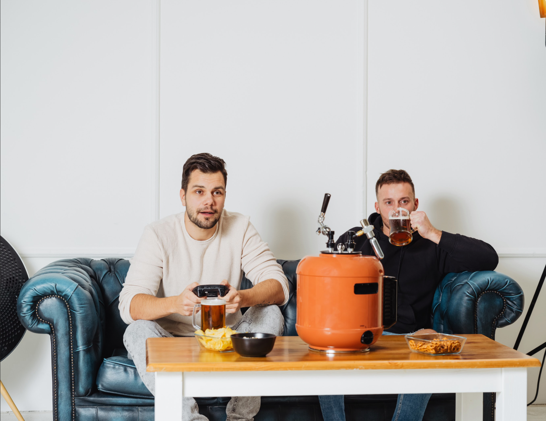Zwei Männer sitzen auf einem blauen Sofa, einer hält einen Gamecontroller, der andere Bier.
