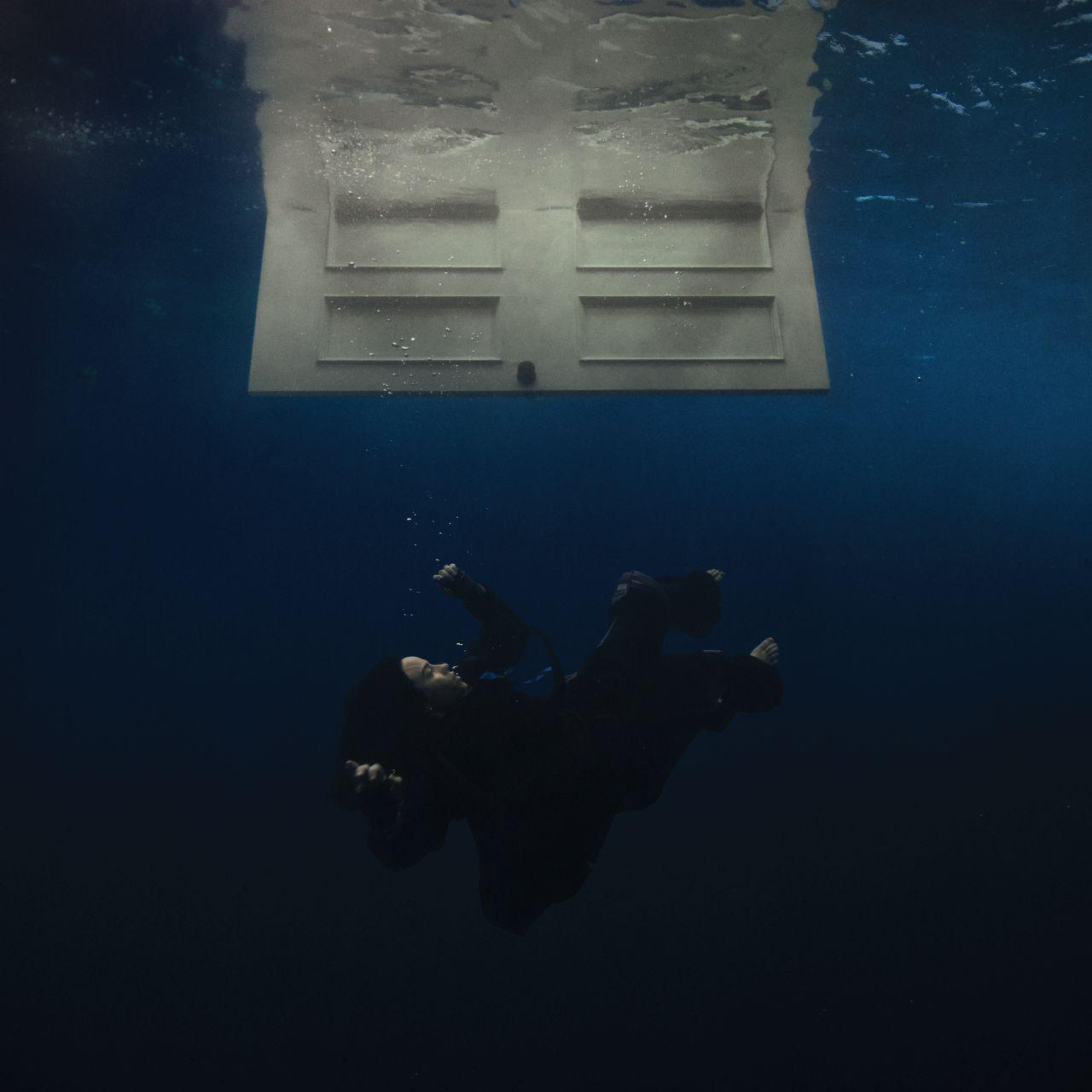 Eine Person unter Wasser, mit einer Tür darüber. Die Person fällt, dunkle Kleidung.