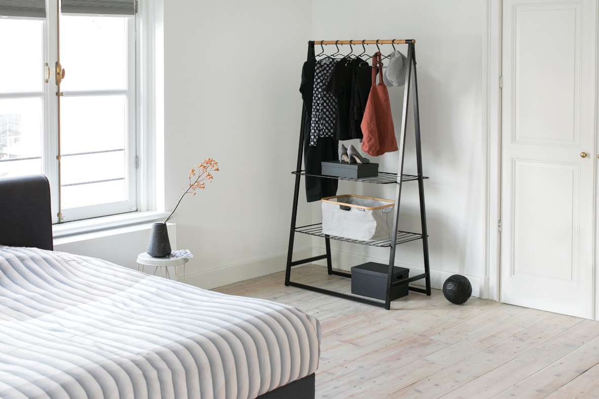 Schlafzimmer mit Kleiderständer. Fenster, Bett und Tür sichtbar. Weiße Wände und Holzboden.