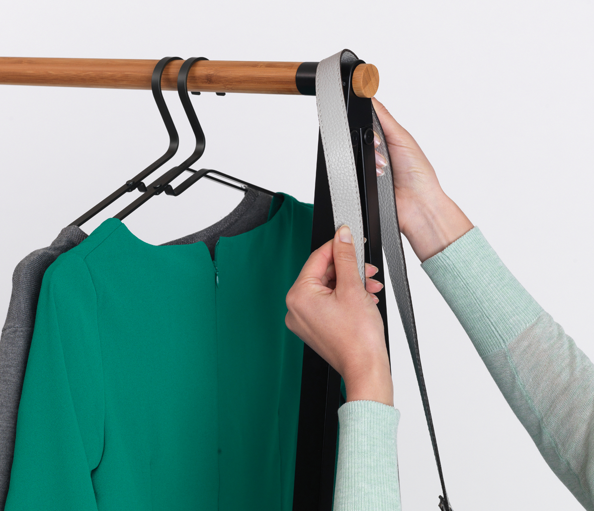Hände hängen einen grauen Riemen an eine Kleiderstange mit grünem Kleid und Kleiderbügeln.
