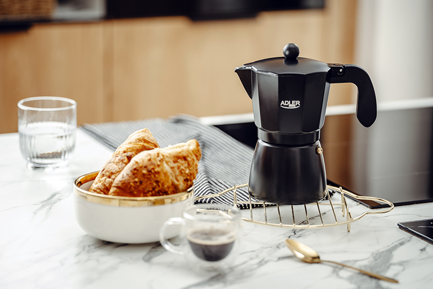 Fekete mokkakávéfőző egy arany alátéten, croissant-ok és kávé mellett.