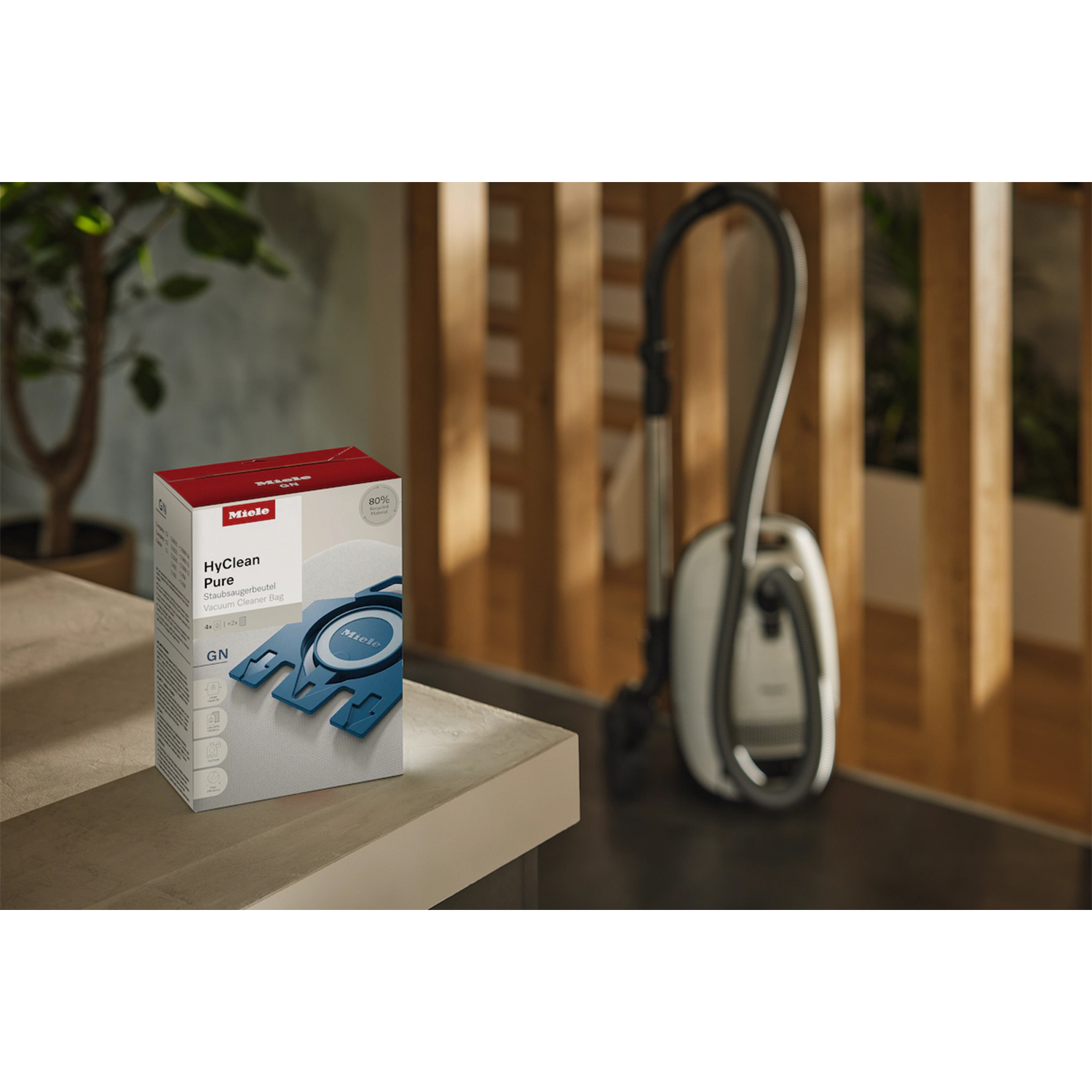 Una scatola di sacchetti per aspirapolvere Miele su un tavolo, con un aspirapolvere sullo sfondo.