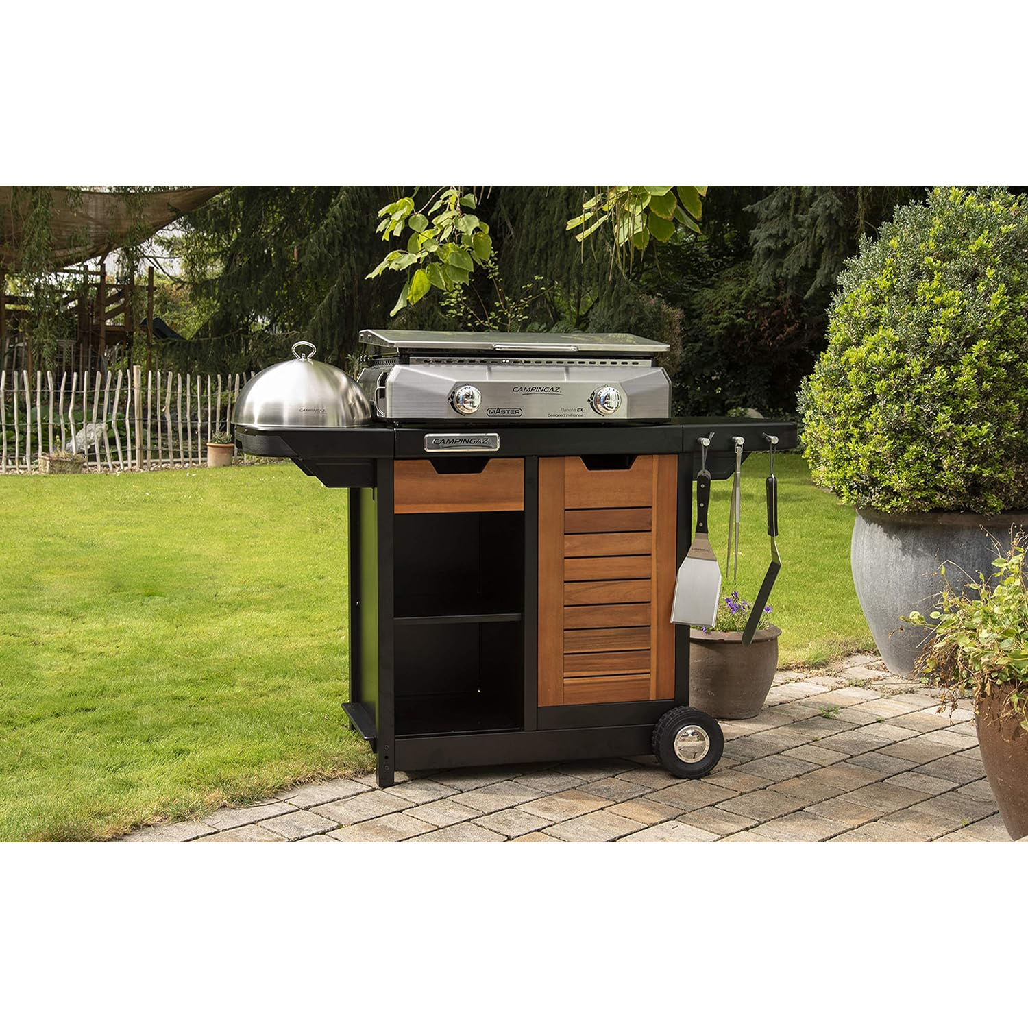 Un barbecue camping gaz su un carrello nero con tavoli laterali e un armadio in legno. Su erba verde.