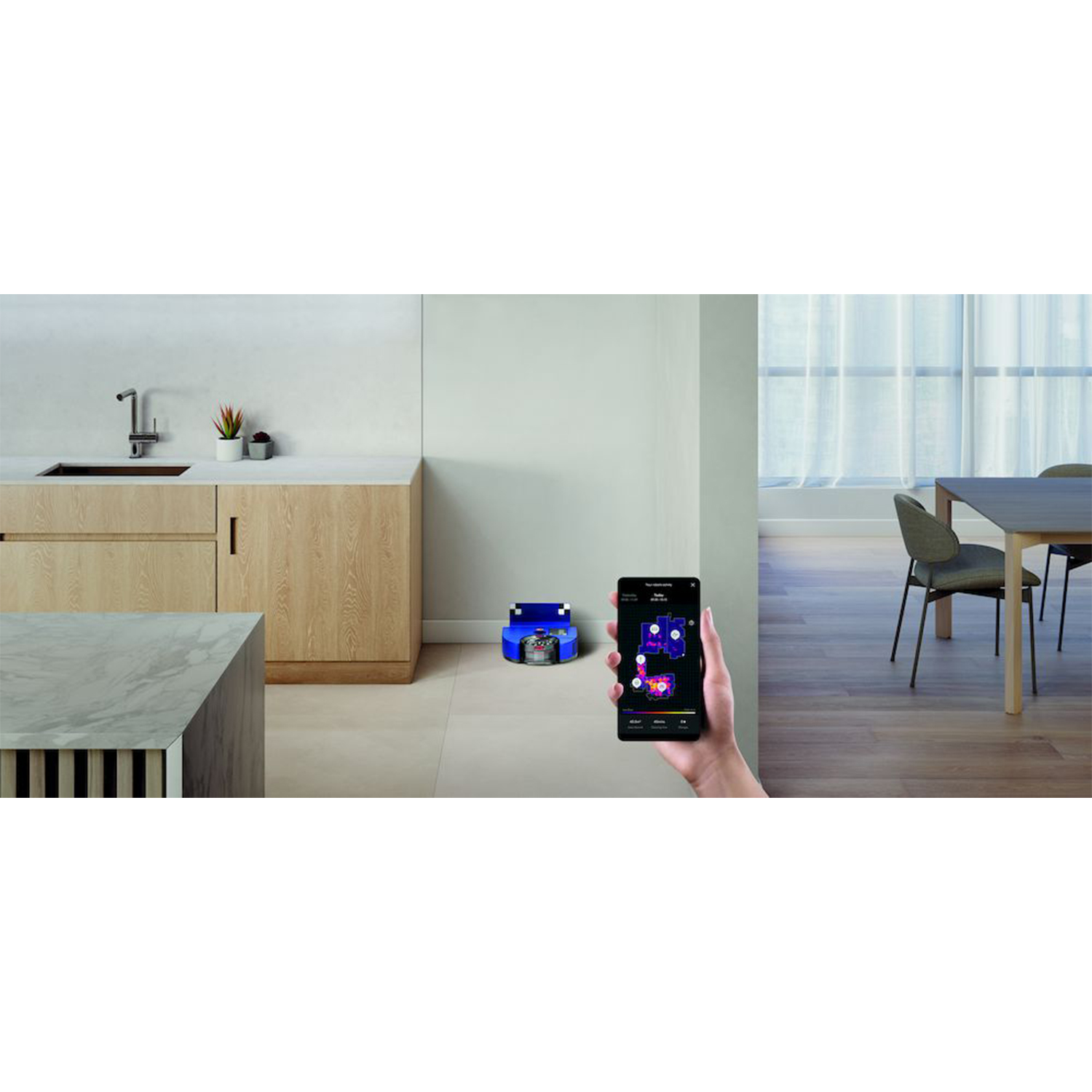 Stanza con aspirapolvere robot. Una mano tiene uno smartphone, lo sfondo è una cucina.