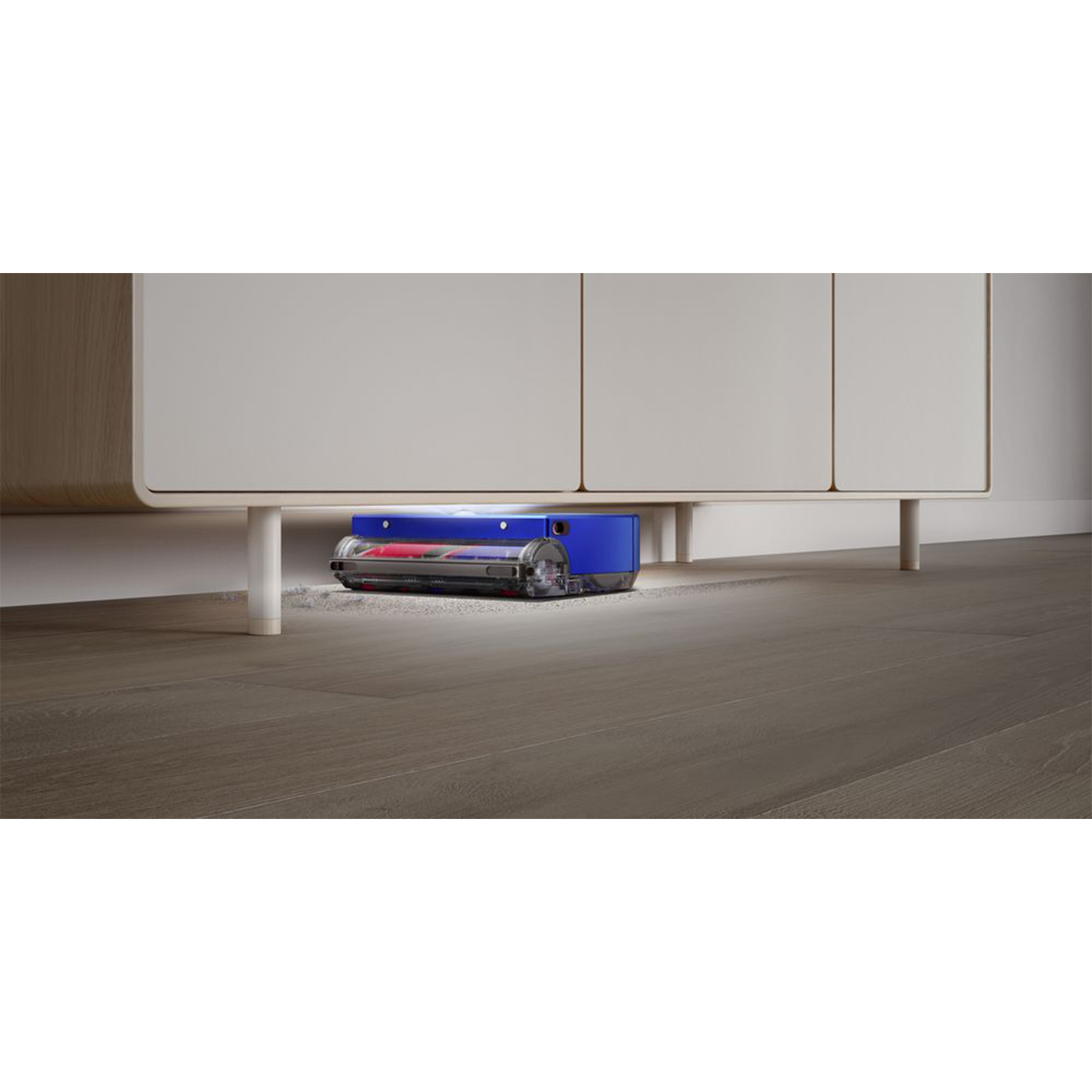 Un aspirapolvere blu e rosso è sotto un mobile bianco. Sta pulendo un pavimento in legno marrone chiaro.