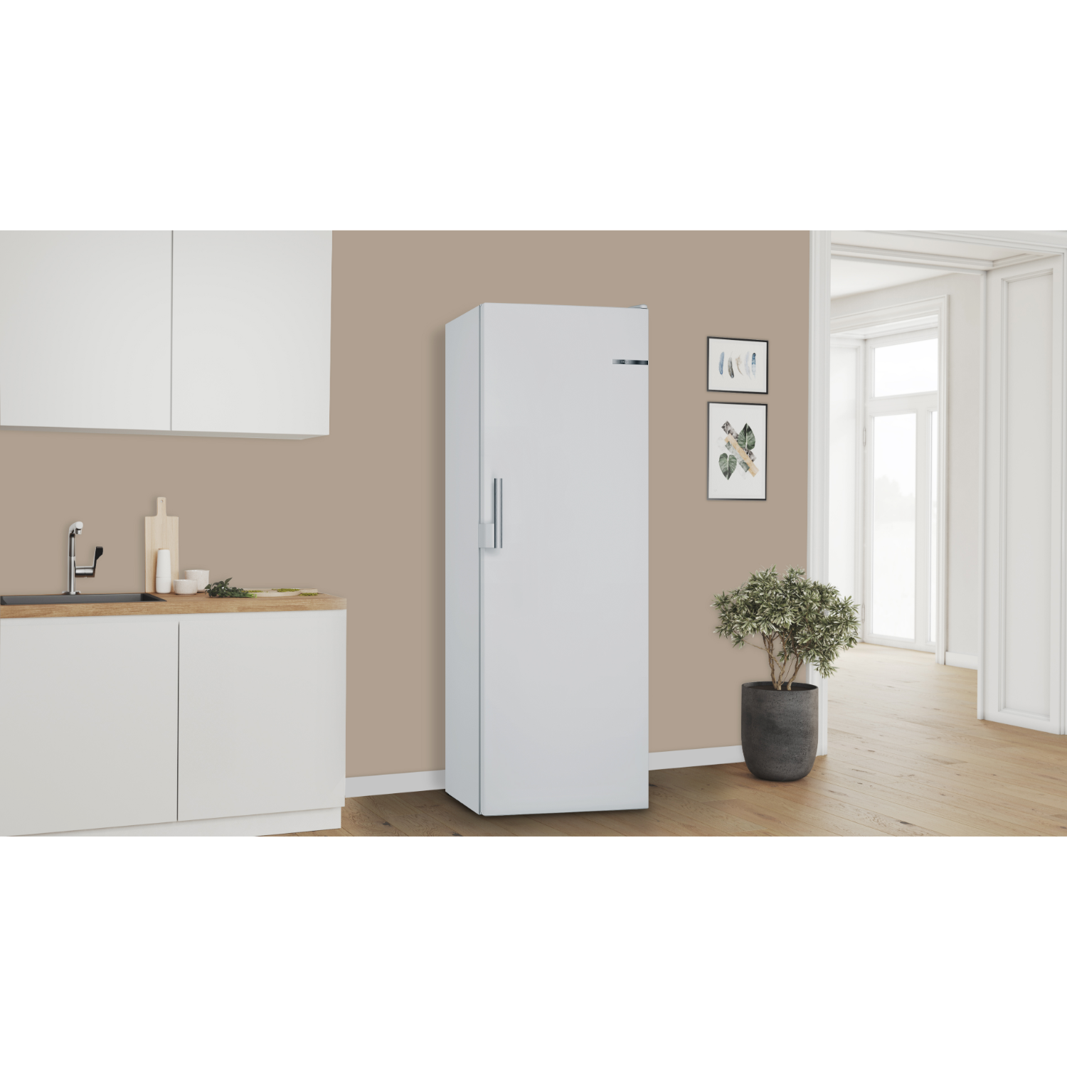 A white refrigerator standing in a kitchen. It has a handle, and a wall is behind it with pictures.