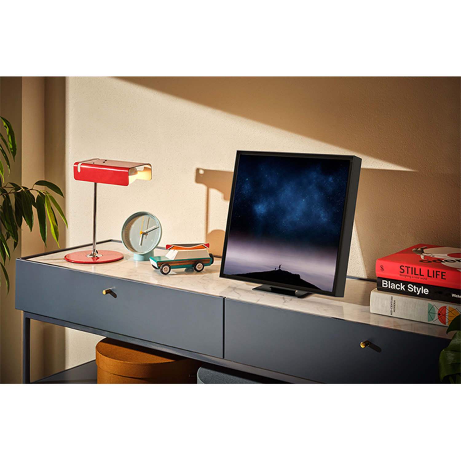 Un bureau avec un moniteur, une lampe, une horloge, une voiture miniature et des livres.