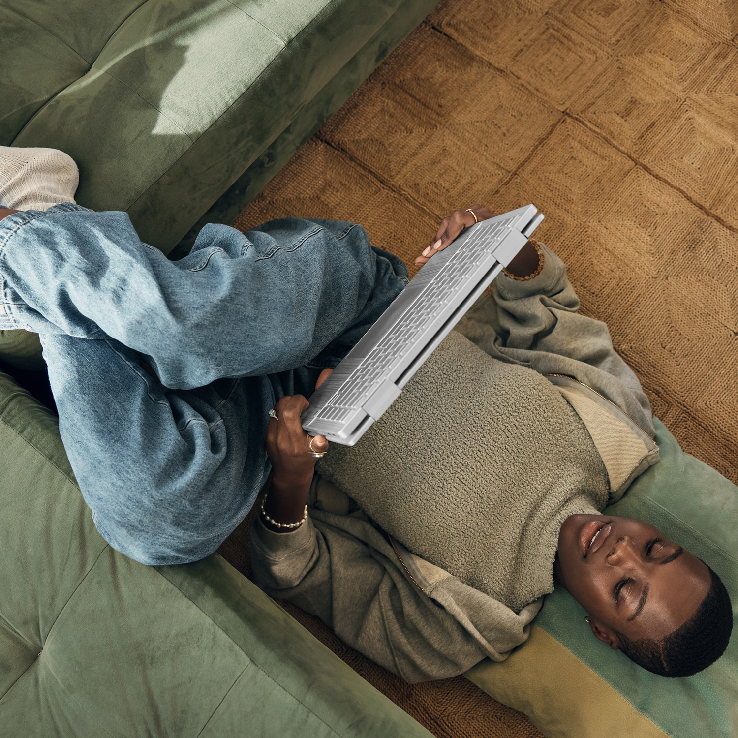 Een persoon die op een groene bank ligt, een laptop gebruikt, een spijkerbroek en een grijze hoodie draagt.