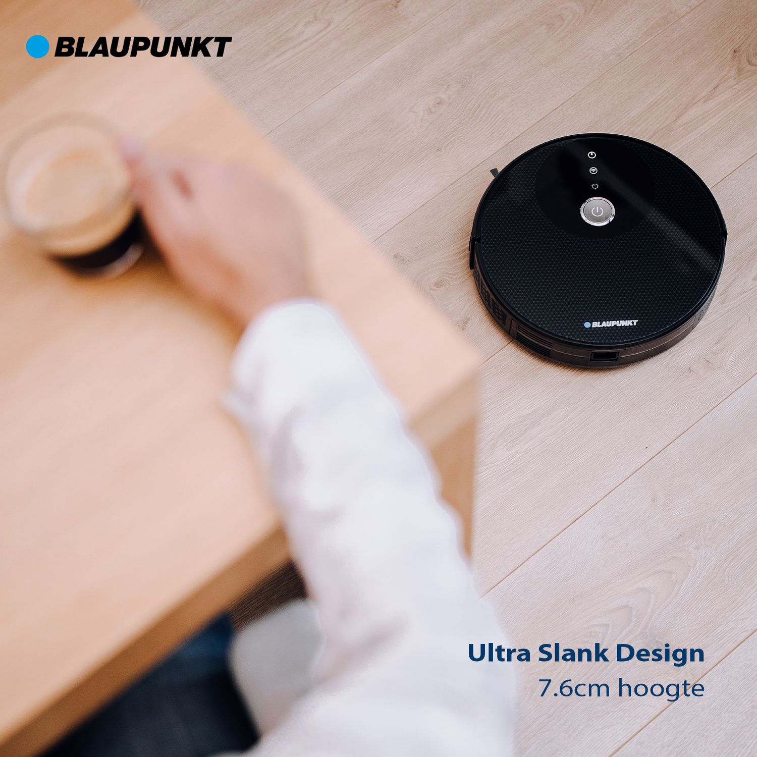 Een zwarte robotstofzuiger op houten vloer. Een hand met een glas koffie is links.