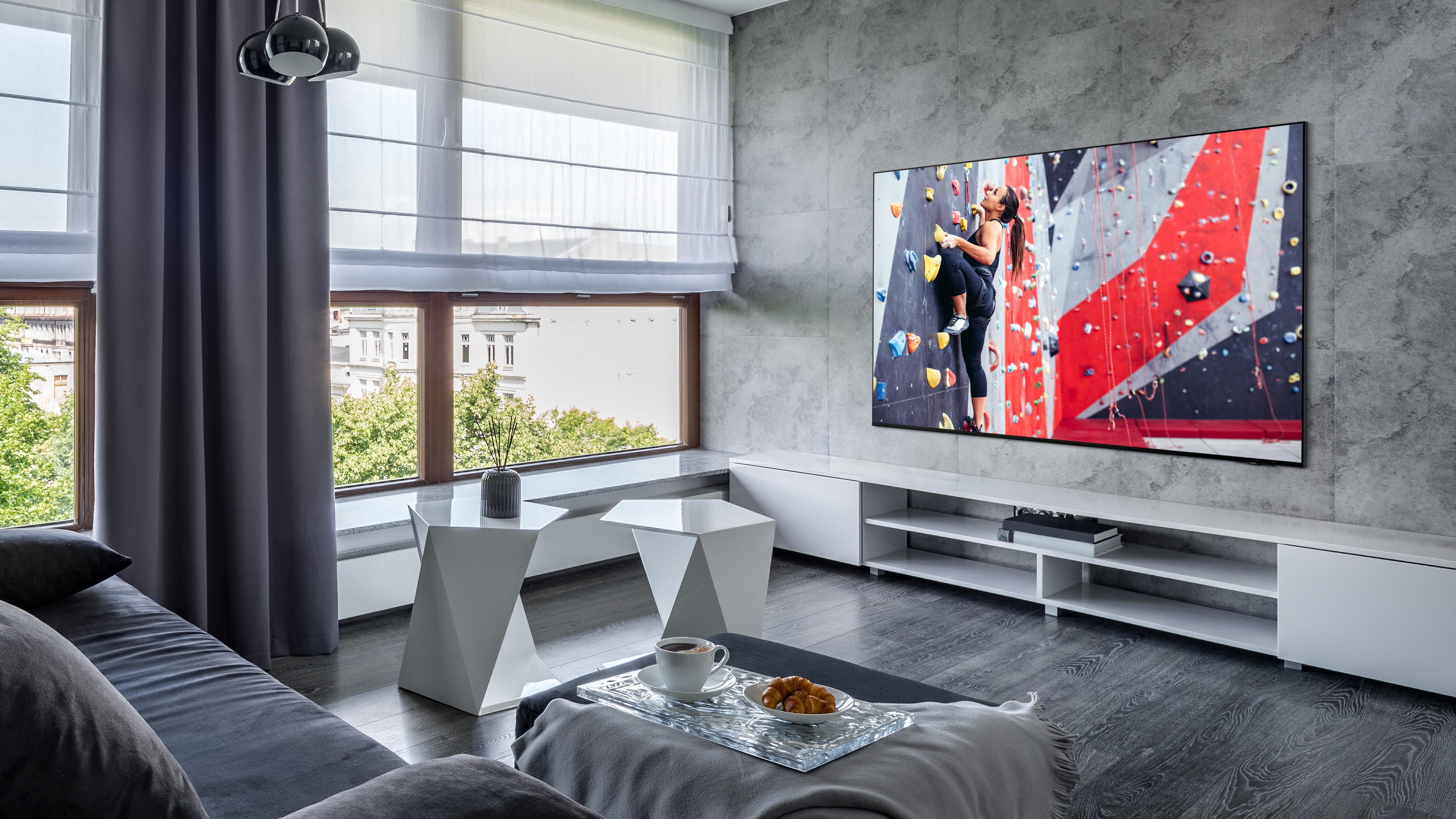 Wohnzimmer mit großem TV, Kletterszene, weißen Tischen und grauem Sofa.