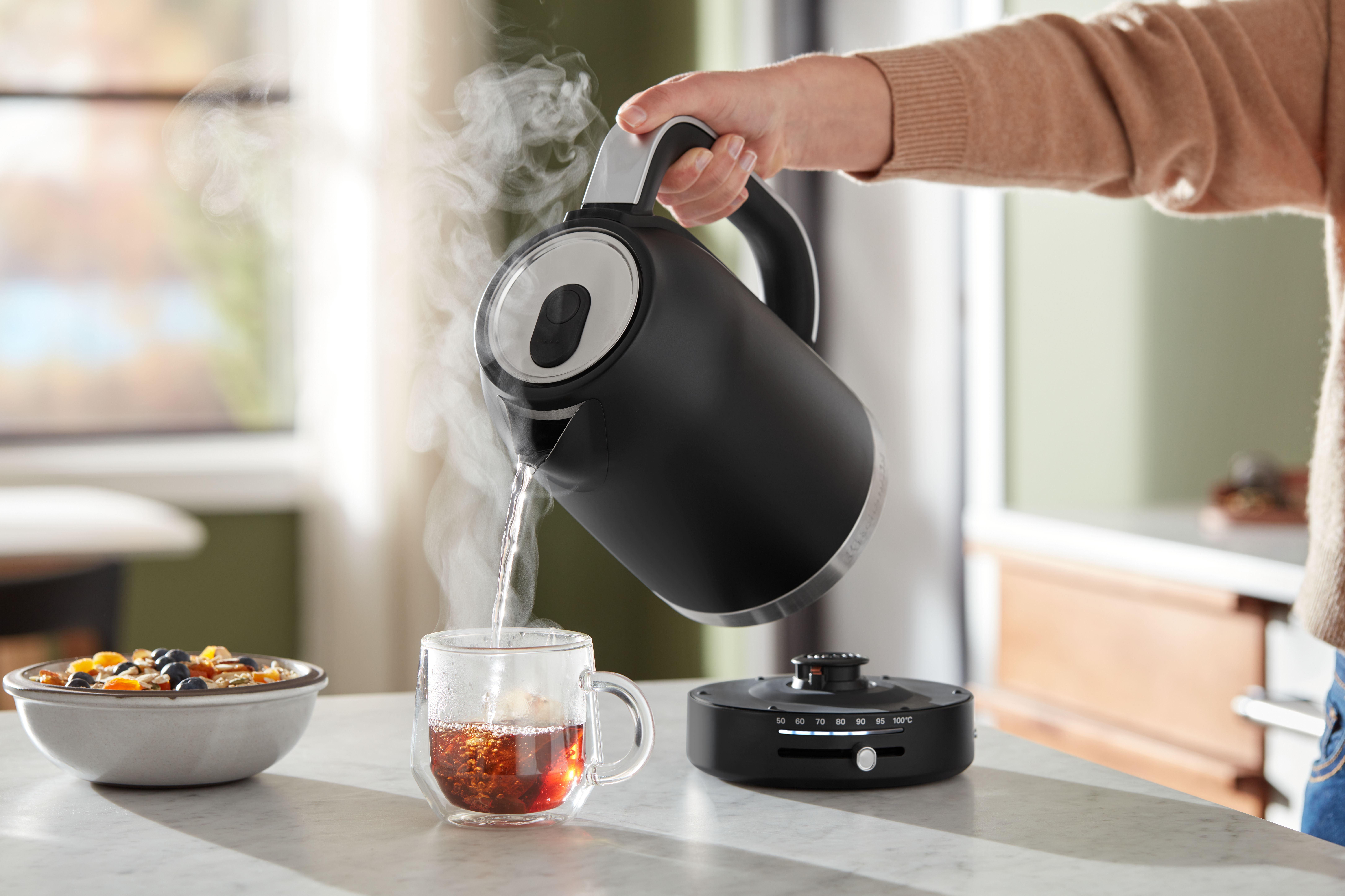 Eine Person gießt heißes Wasser aus einem schwarzen Wasserkocher in eine Tasse.