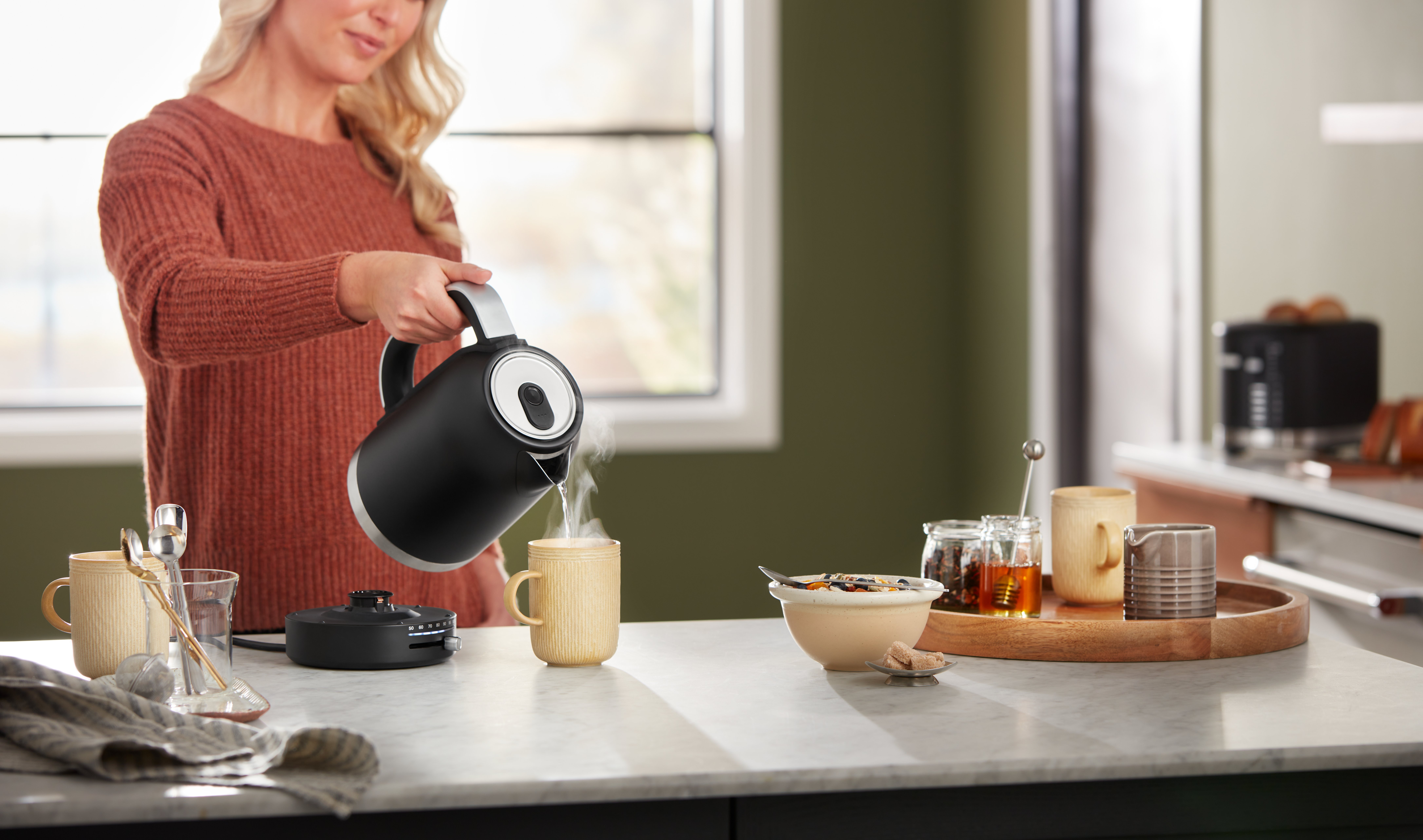 Frau gießt heißes Wasser aus einem schwarzen Wasserkocher in eine Tasse. Frühstücksartikel sind im Hintergrund.