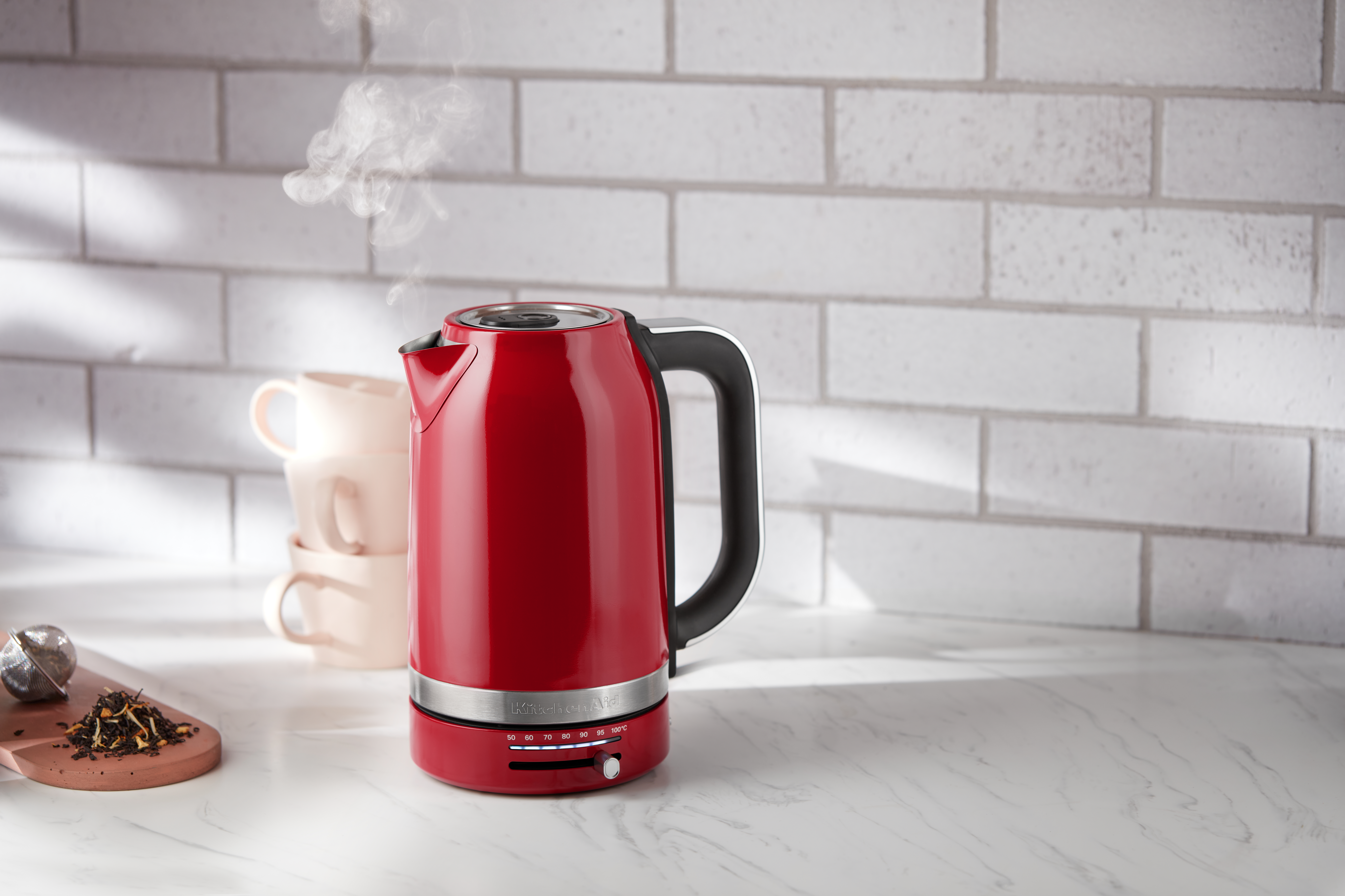 Roter Wasserkocher mit schwarzem Griff, Dampf steigt auf, auf weißer Arbeitsplatte.