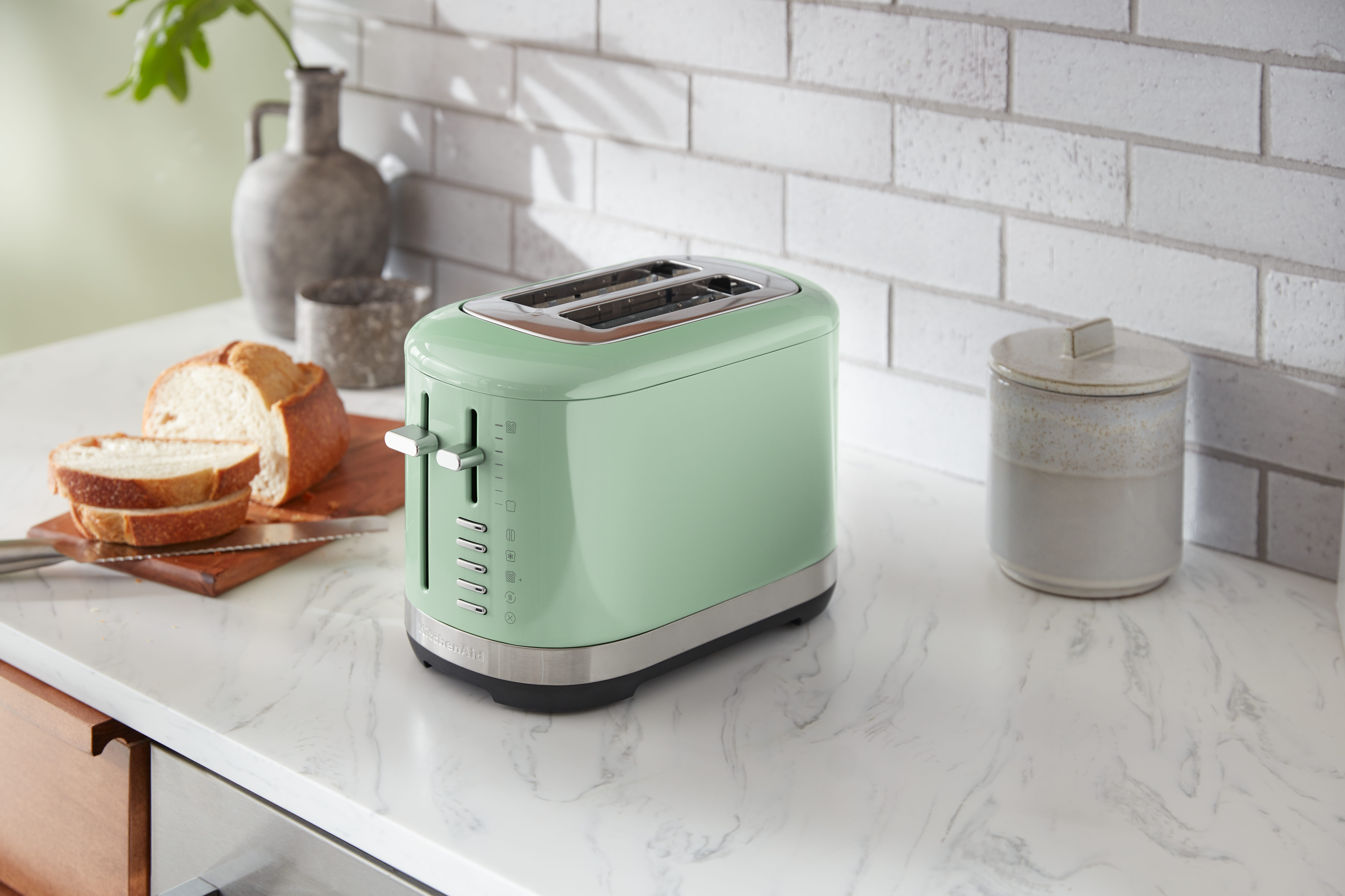 Grüner Toaster mit Brotscheiben auf einem Holzbrett. Ein Glas und eine Vase im Hintergrund.