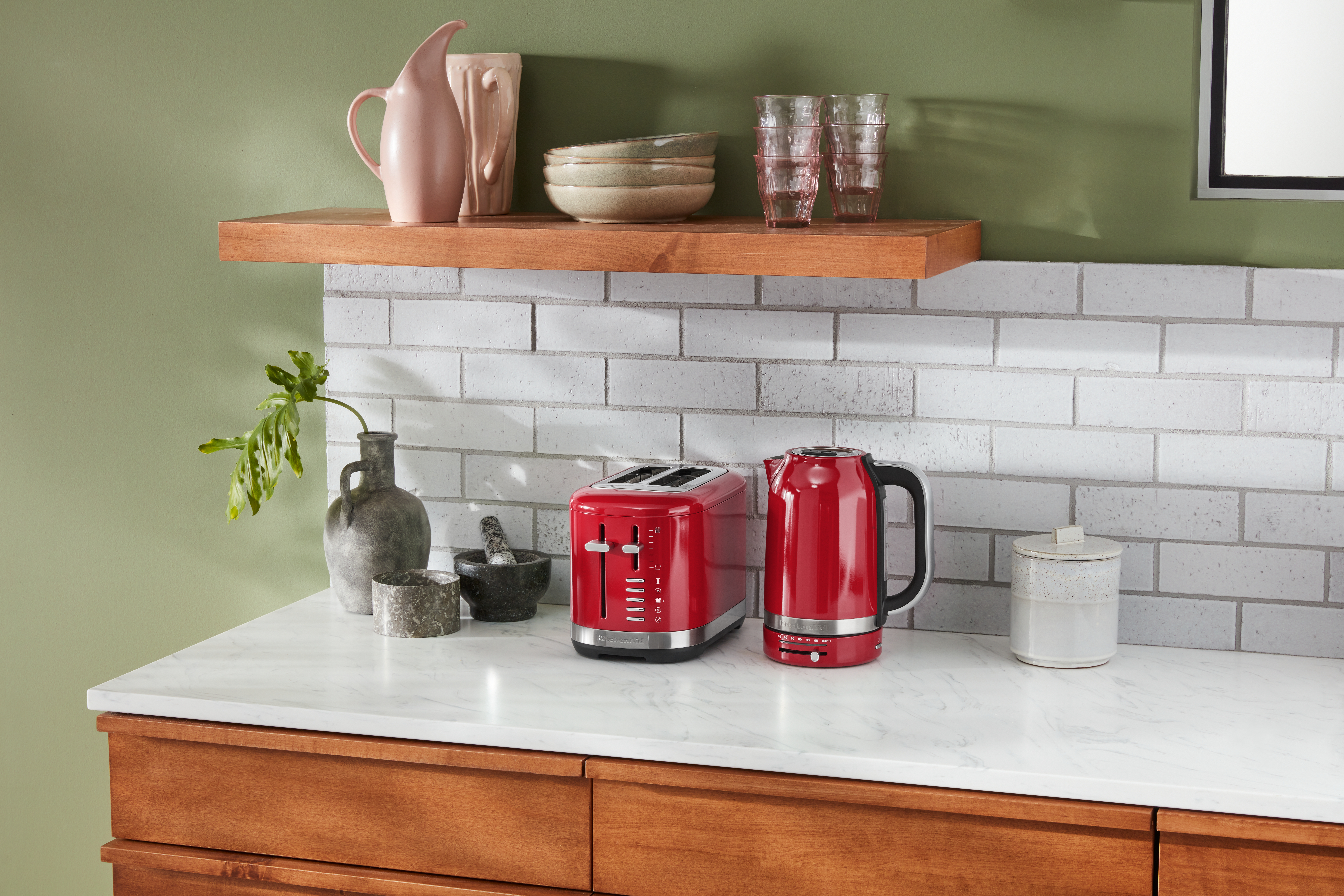 Küchenzeile mit rotem Toaster, Wasserkocher und Keramikartikeln vor einer Ziegelmauer und grünem Hintergrund.