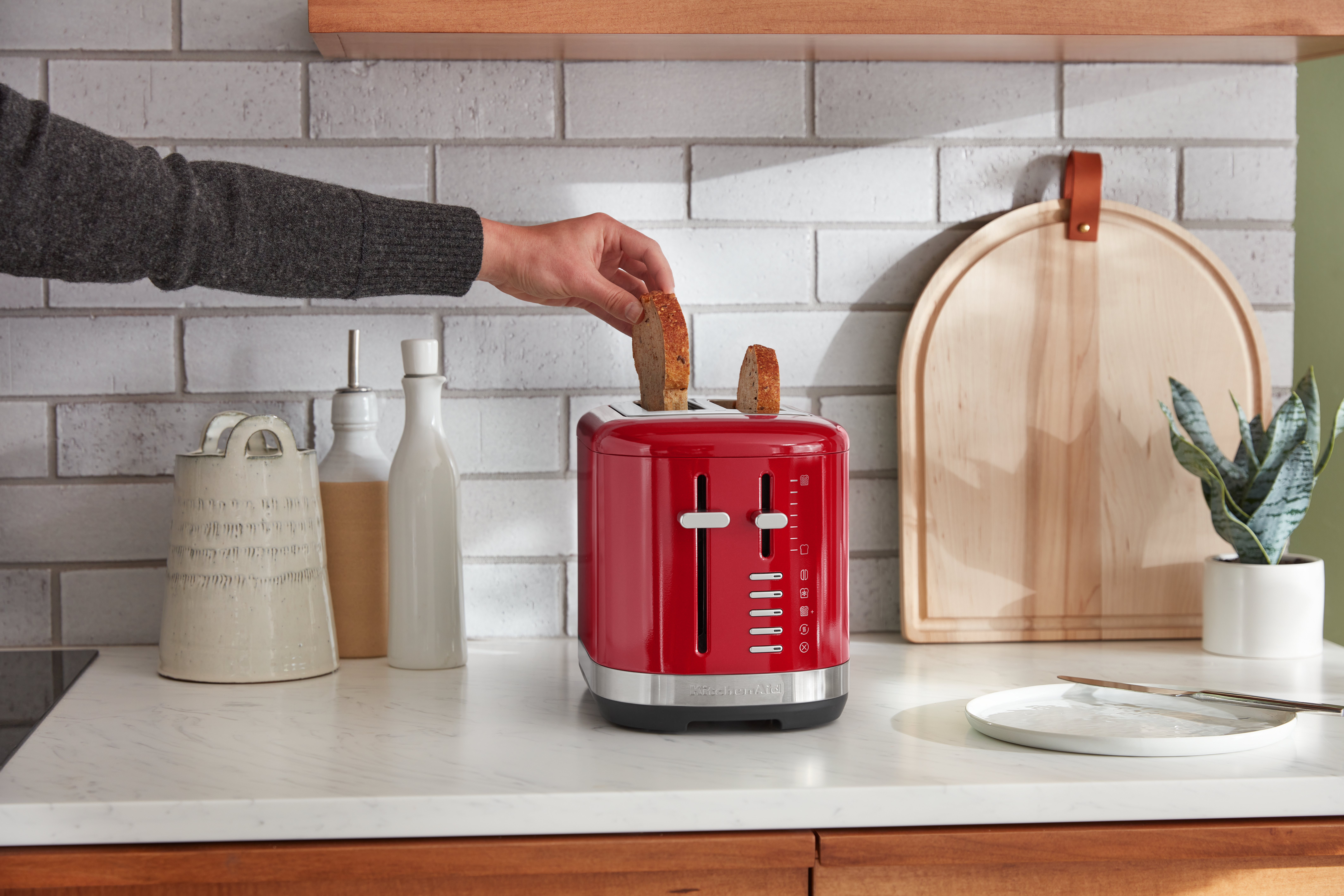Eine Hand legt Brot in einen roten KitchenAid-Toaster auf einer weißen Arbeitsplatte mit Gegenständen.