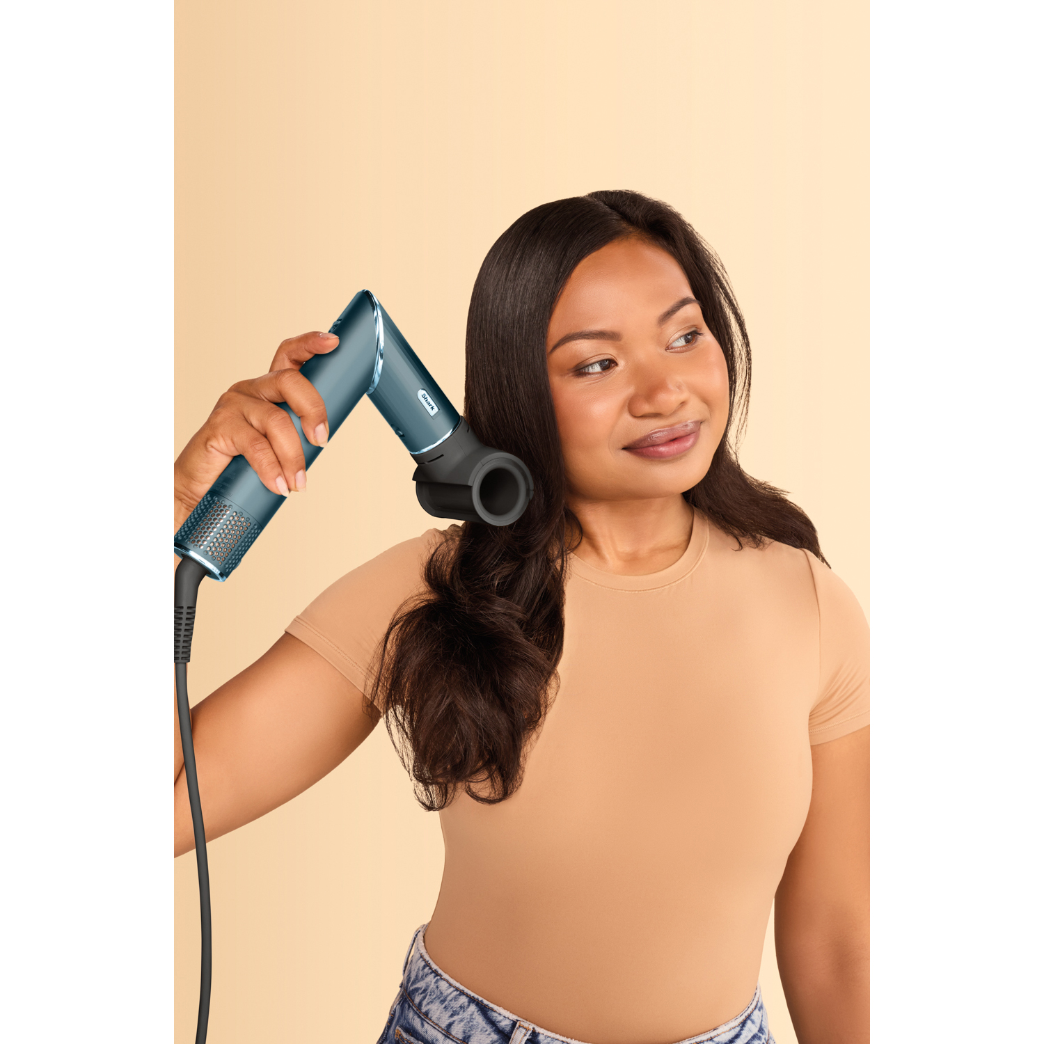 Una donna usa un asciugacapelli verde acqua, asciugando i suoi capelli scuri. Sorride.
