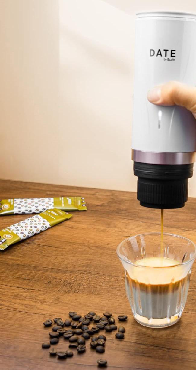 Eine Person gießt Kaffee aus einem weißen tragbaren Espressomaschine in ein Glas, Kaffeebohnen in der Nähe.