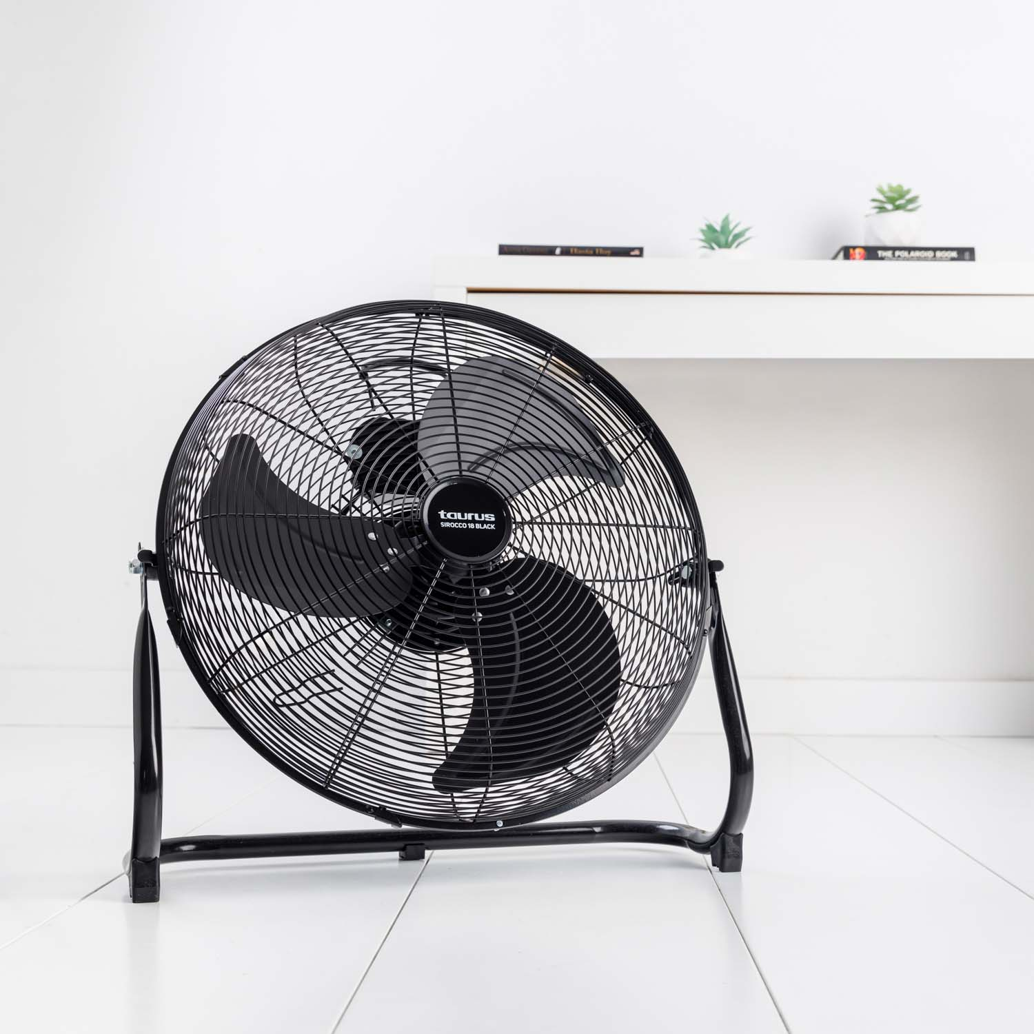 Un ventilador eléctrico negro en un suelo blanco. Fondo: pared blanca y estante con plantas y libros.