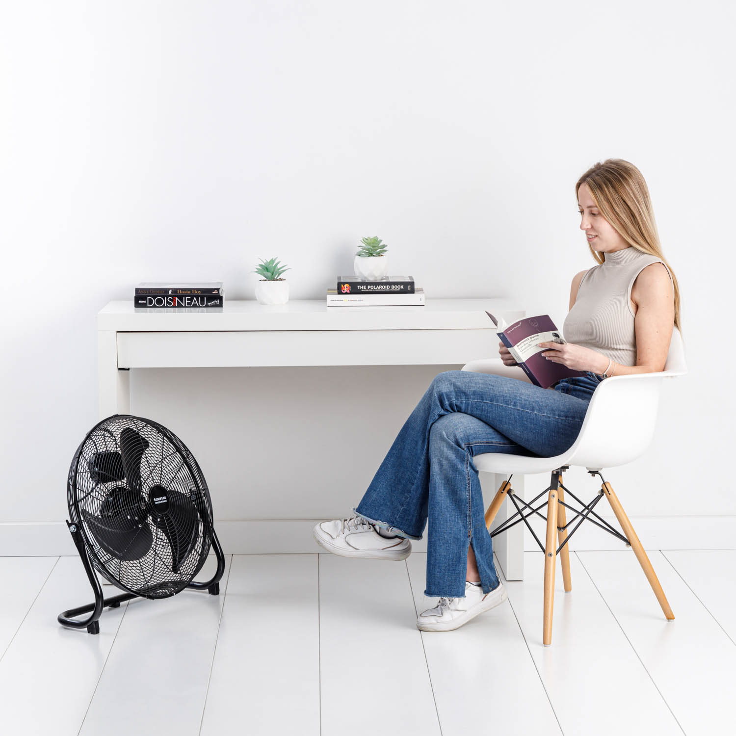 Mujer leyendo un libro con un ventilador, plantas y un escritorio blanco. Suelo blanco.