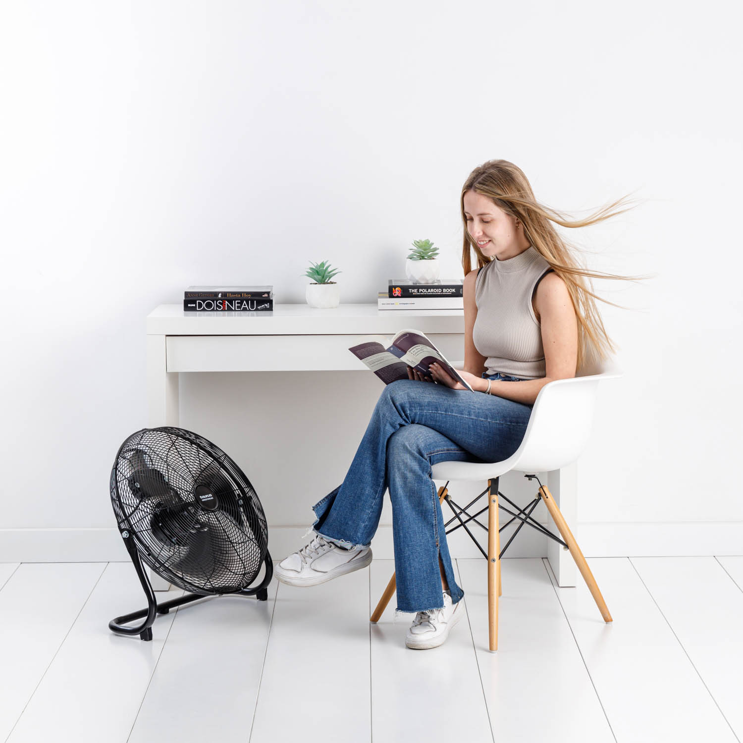 Una mujer leyendo un libro cerca de un ventilador, sentada en una silla blanca en una habitación blanca.