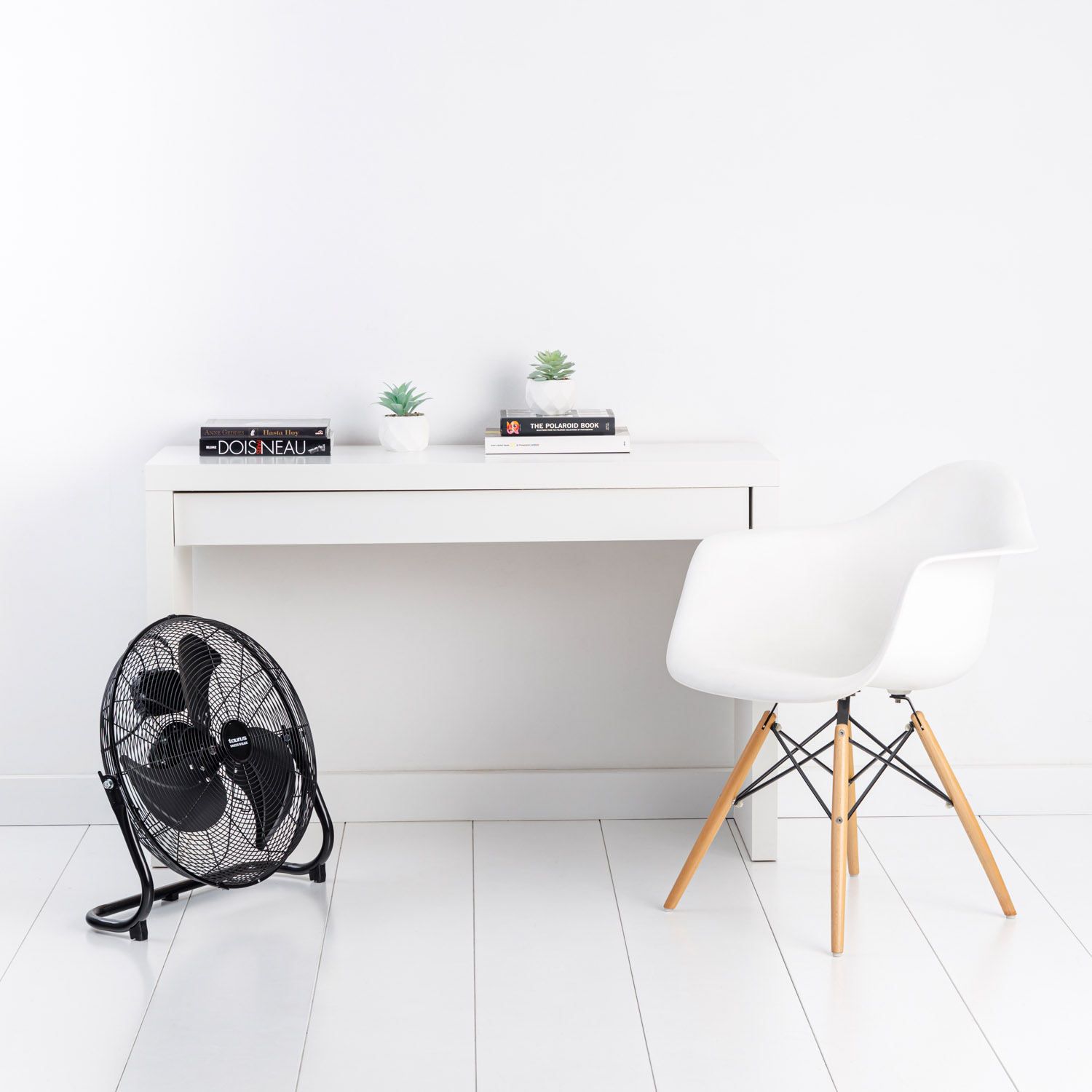 Habitación blanca con escritorio, silla y ventilador negro. Dos plantas en el escritorio.