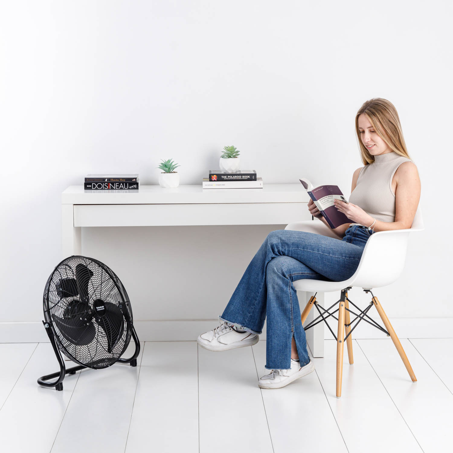 Mujer leyendo un libro, con un ventilador y un escritorio blanco con plantas de fondo.